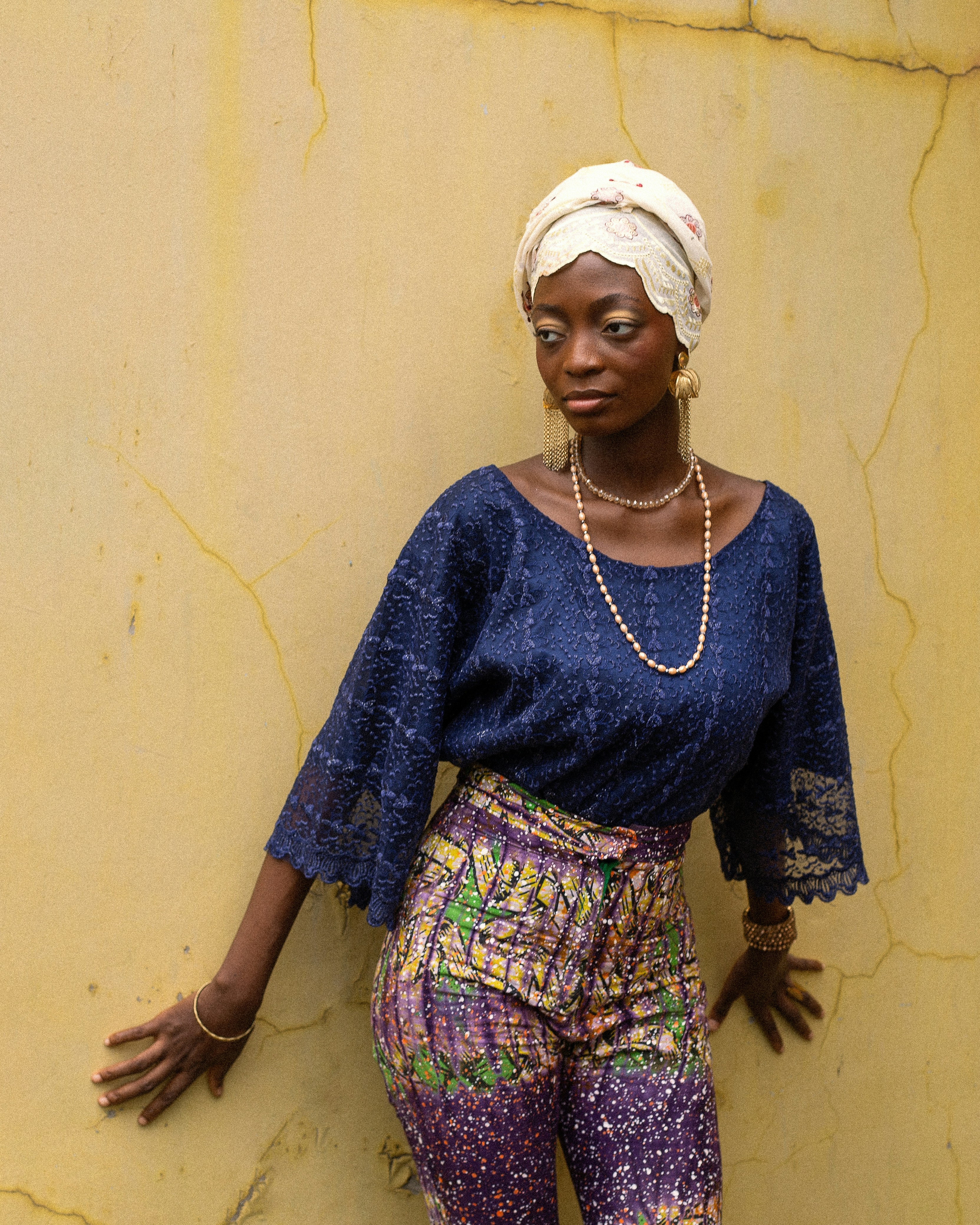 A woman in a blue lace top and patterned trousers against a yellow wall