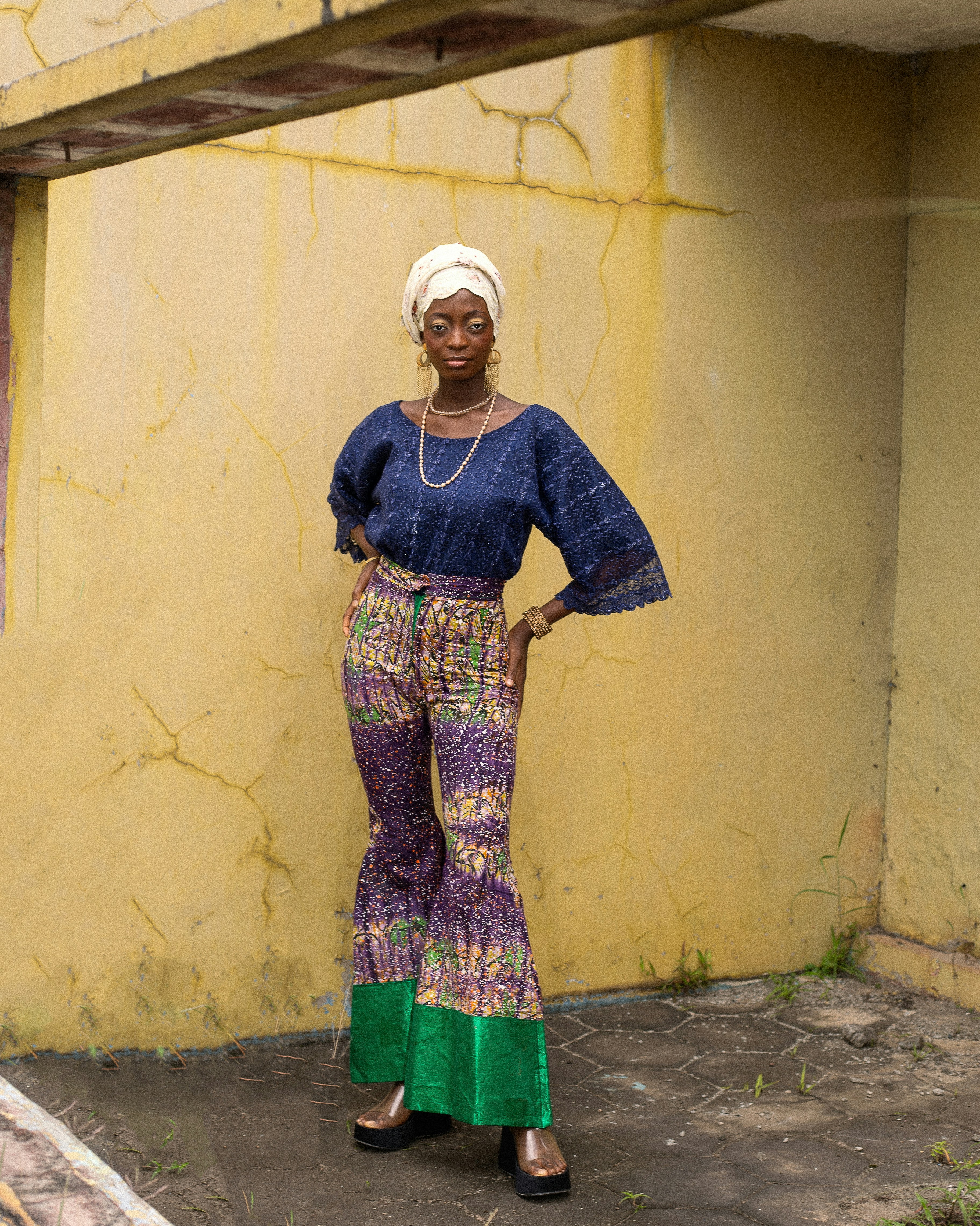 A woman in a blue lace top and patterned flared trousers against a yellow wall