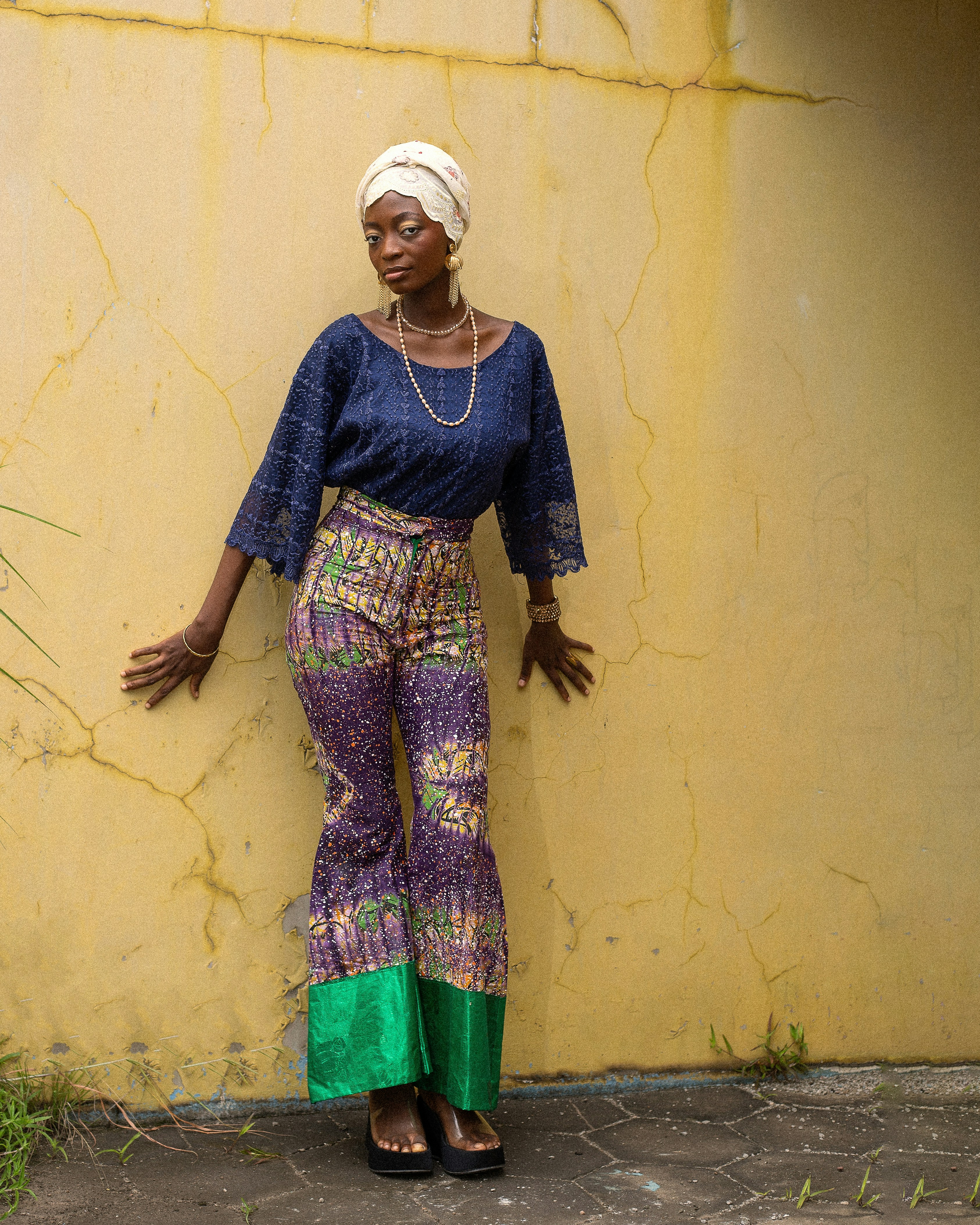 A woman in a blue lace top and patterned flared trousers against a yellow wall