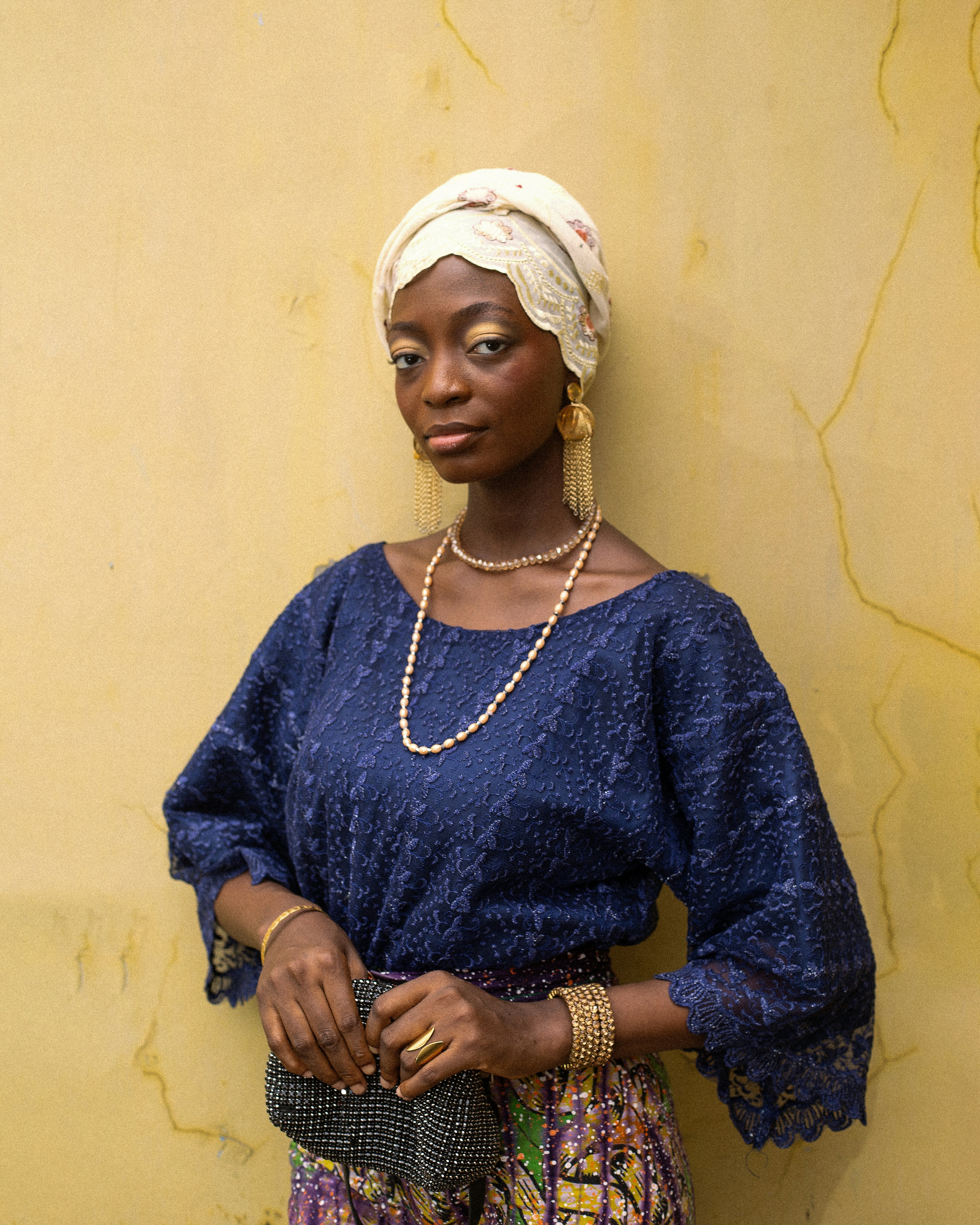 A woman in a blue lace top and headwrap against a yellow wall