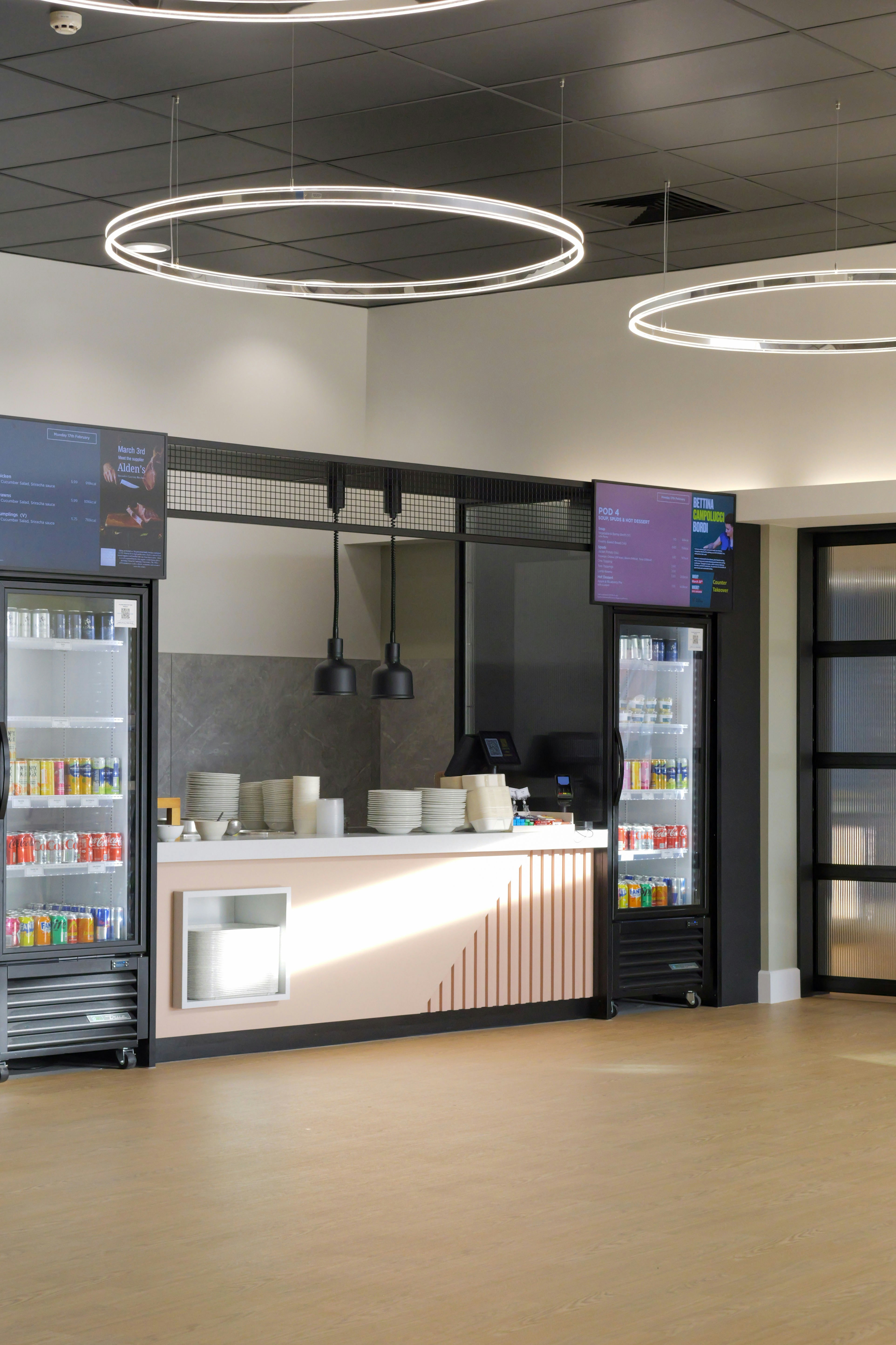 A modern cafeteria counter with glass refrigerators, stacked plates, and circular pendant lights