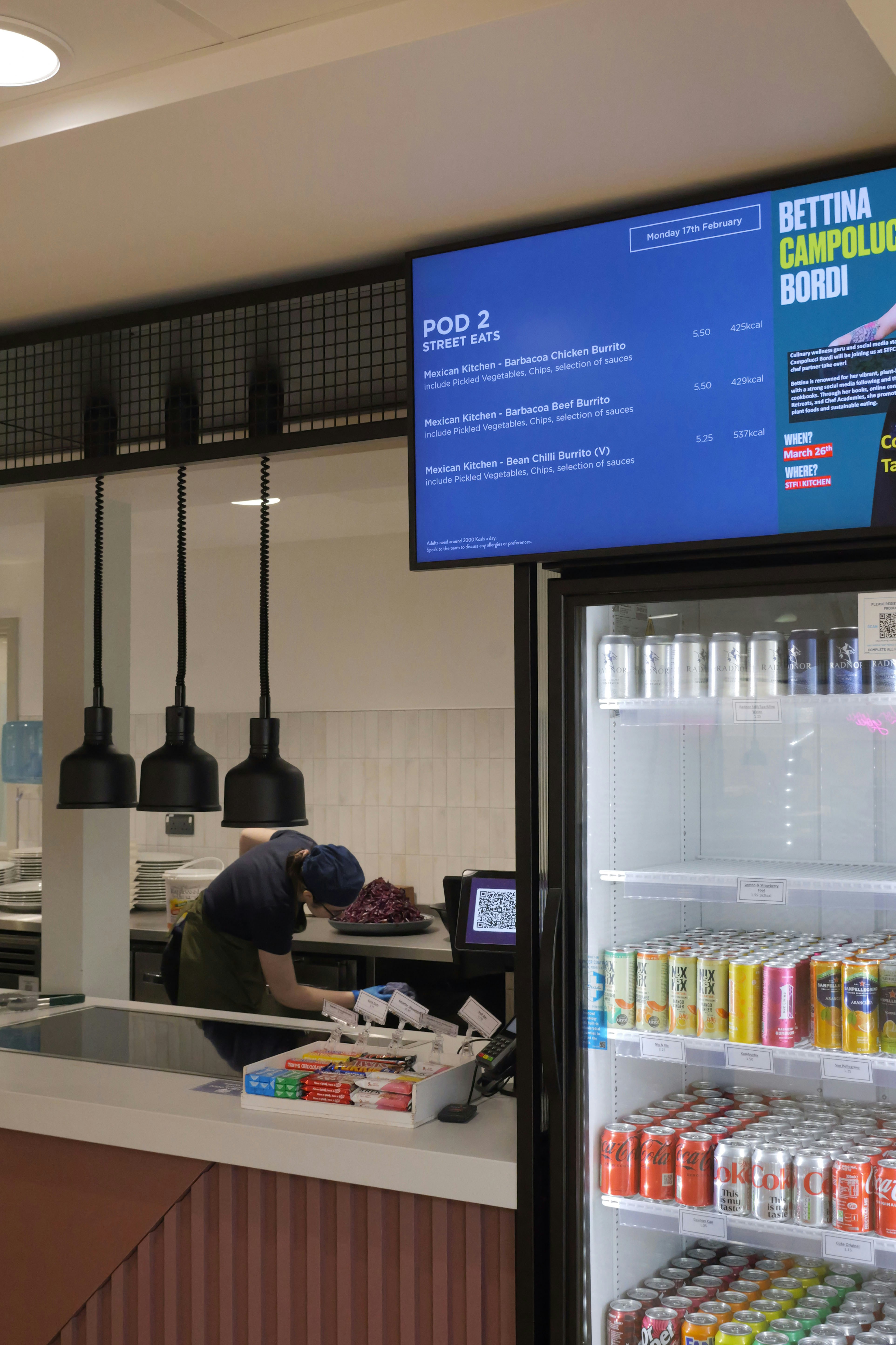 A person in a blue cap and green apron working behind a cafe counter