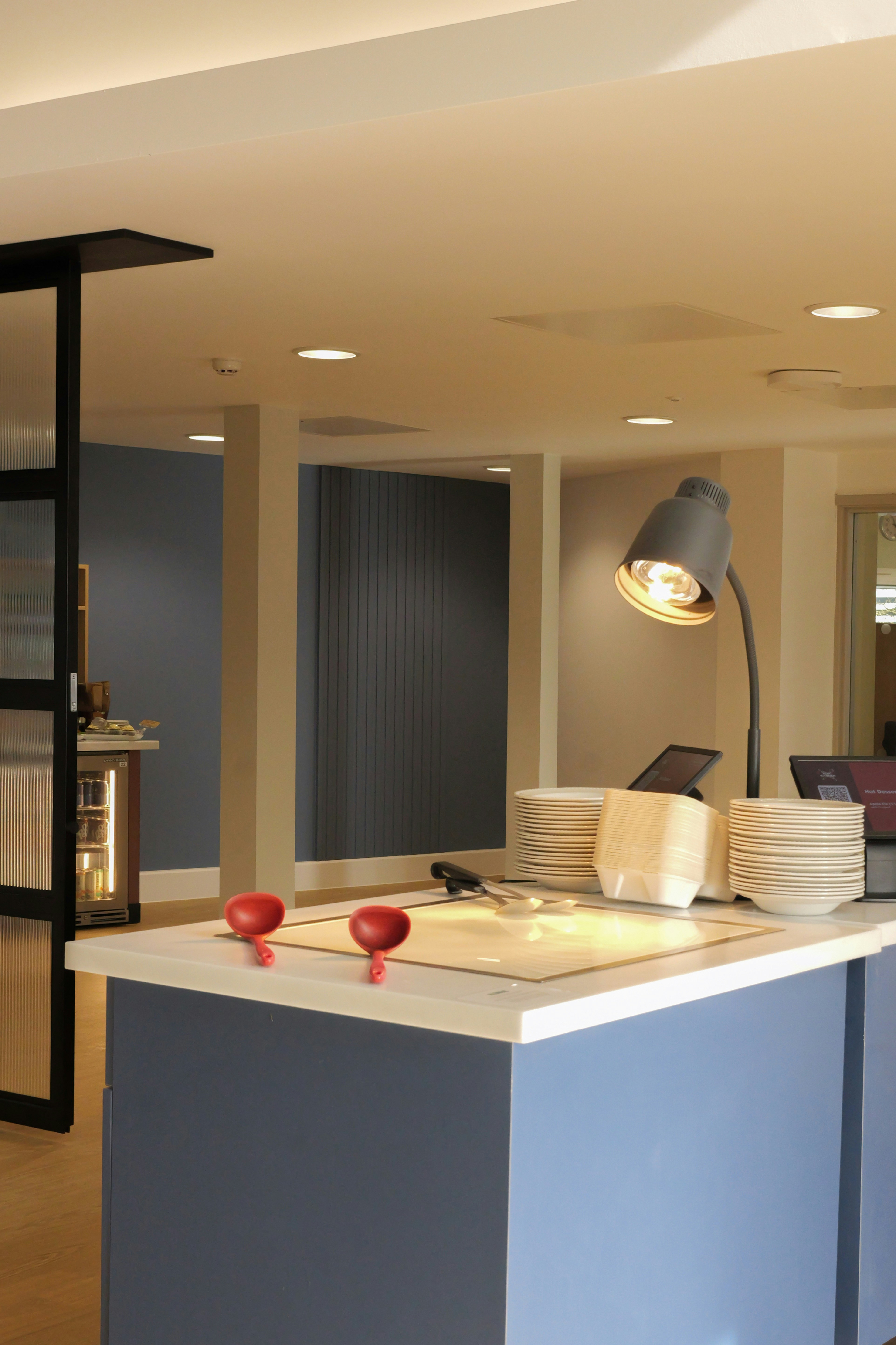 A buffet counter with stacks of plates, red ladles, and a desk lamp