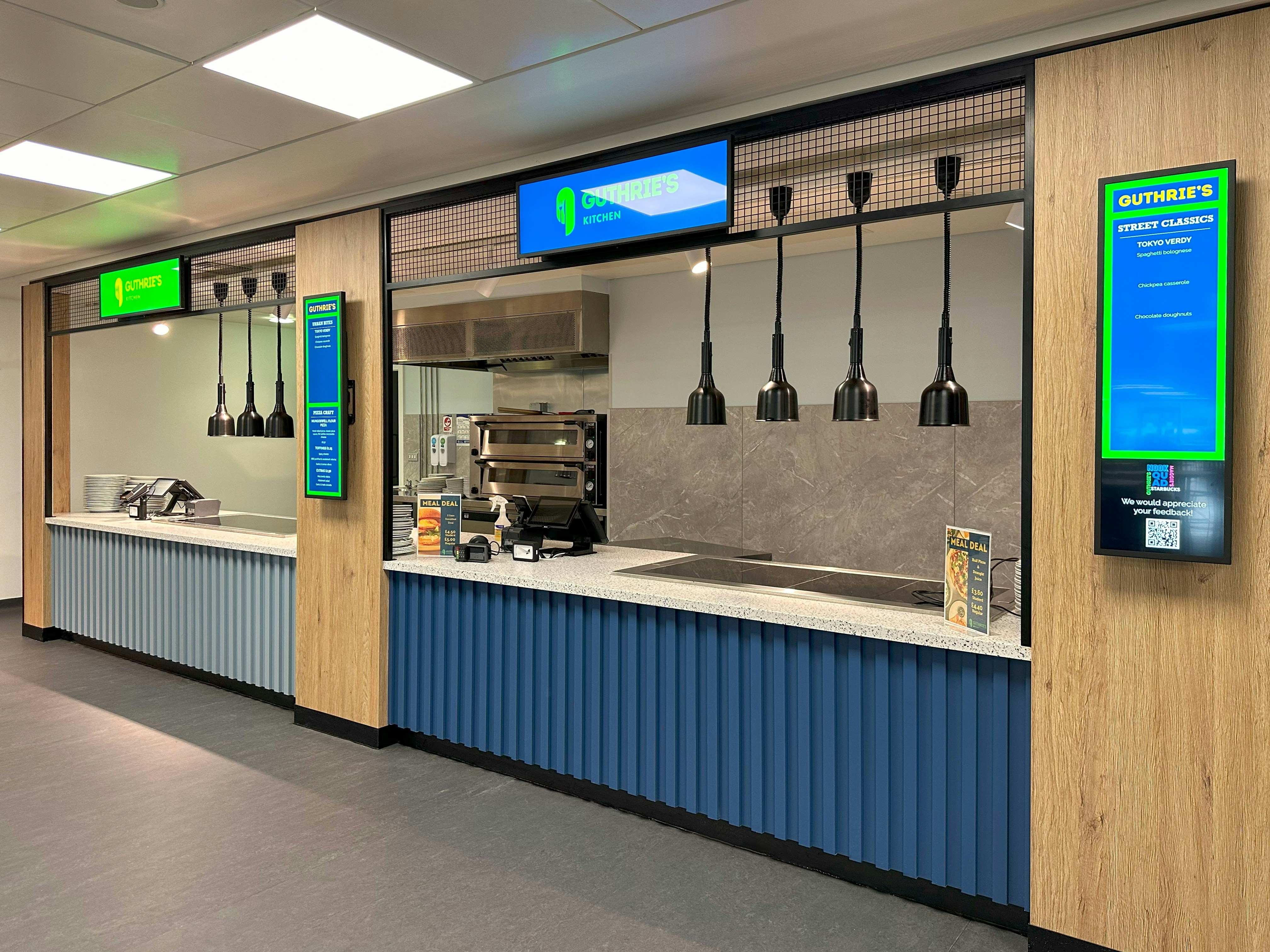 A modern cafeteria serving counter with blue corrugated panels and hanging black pendant lights