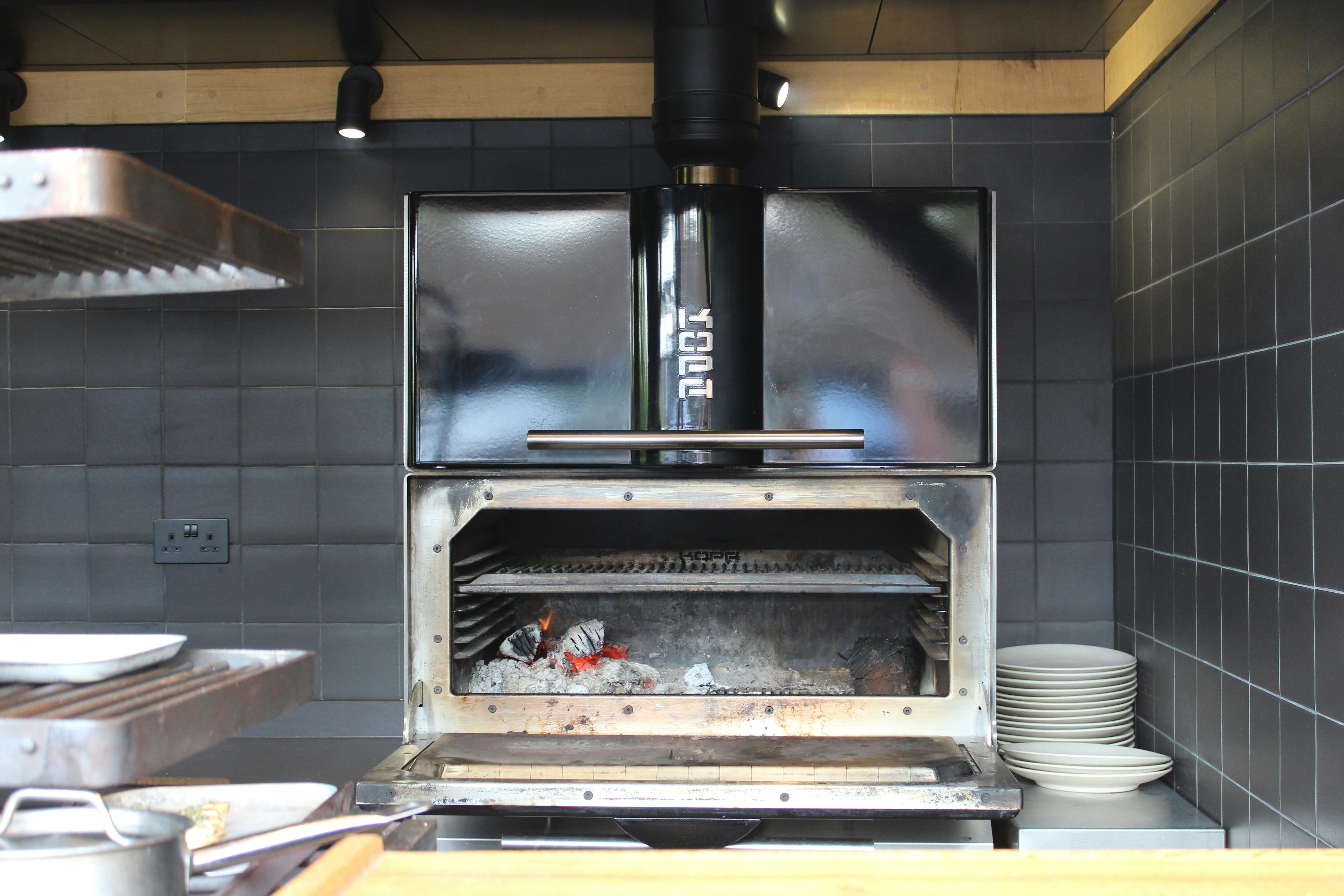 A wood-fired grill with glowing embers inside a black tiled kitchen