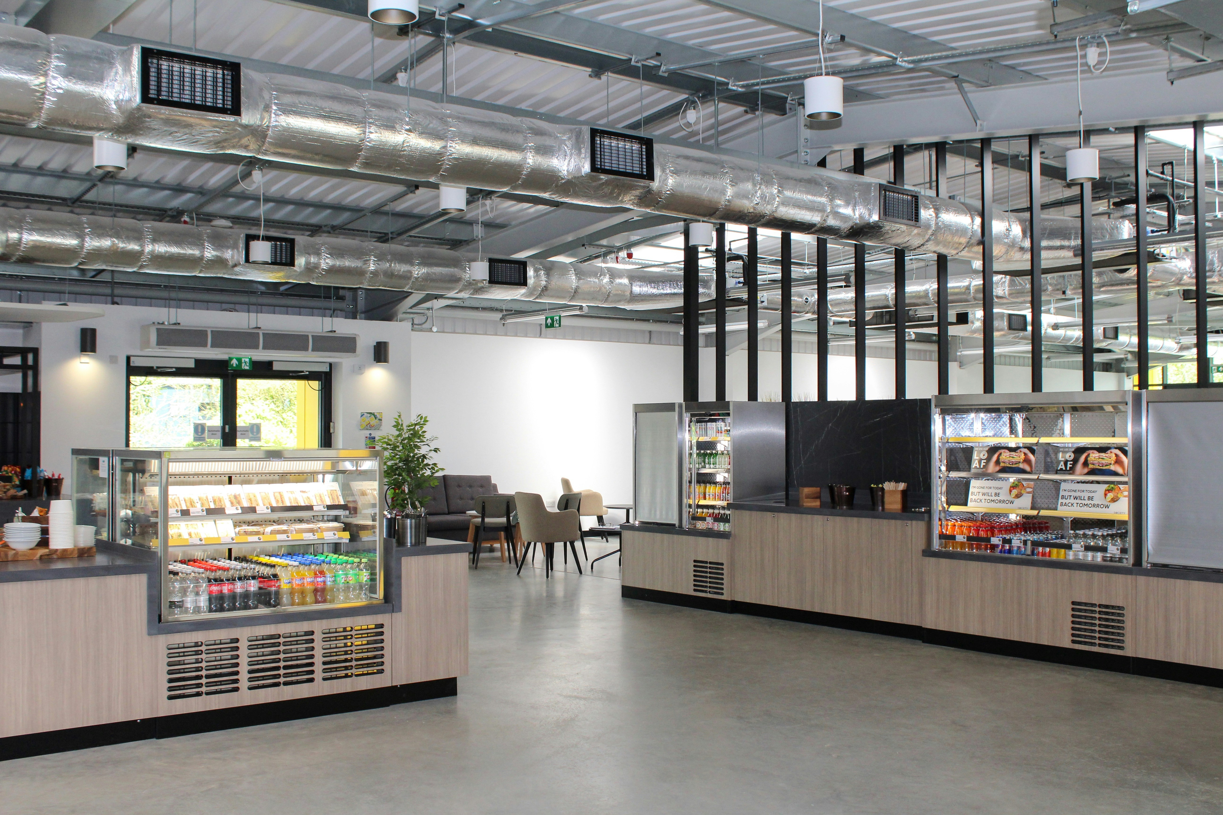 A modern cafeteria with refrigerated display cases, wooden counters, and industrial ceiling ductwork
