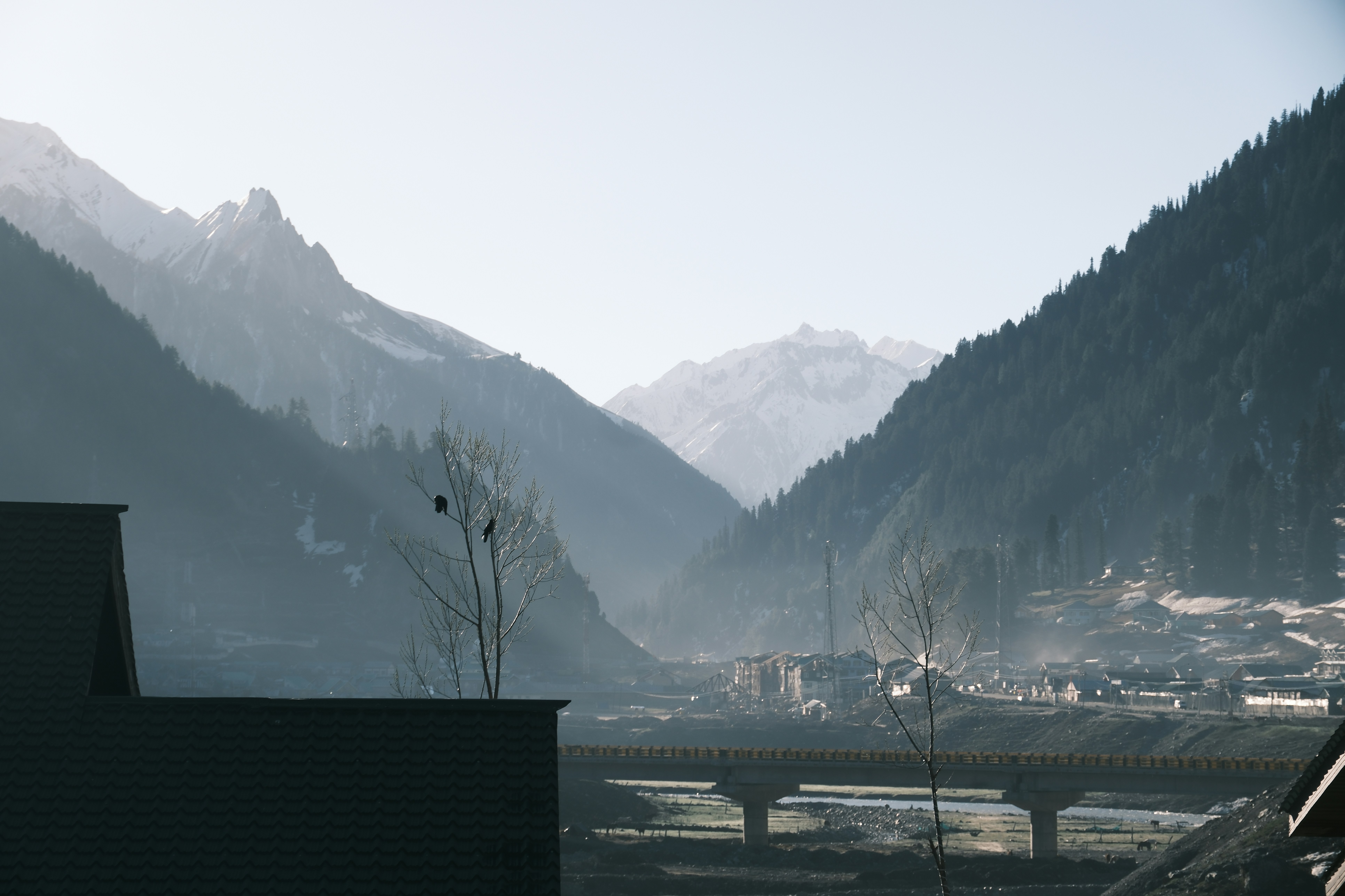 A misty mountain valley with a bridge and small buildings under a bright sky