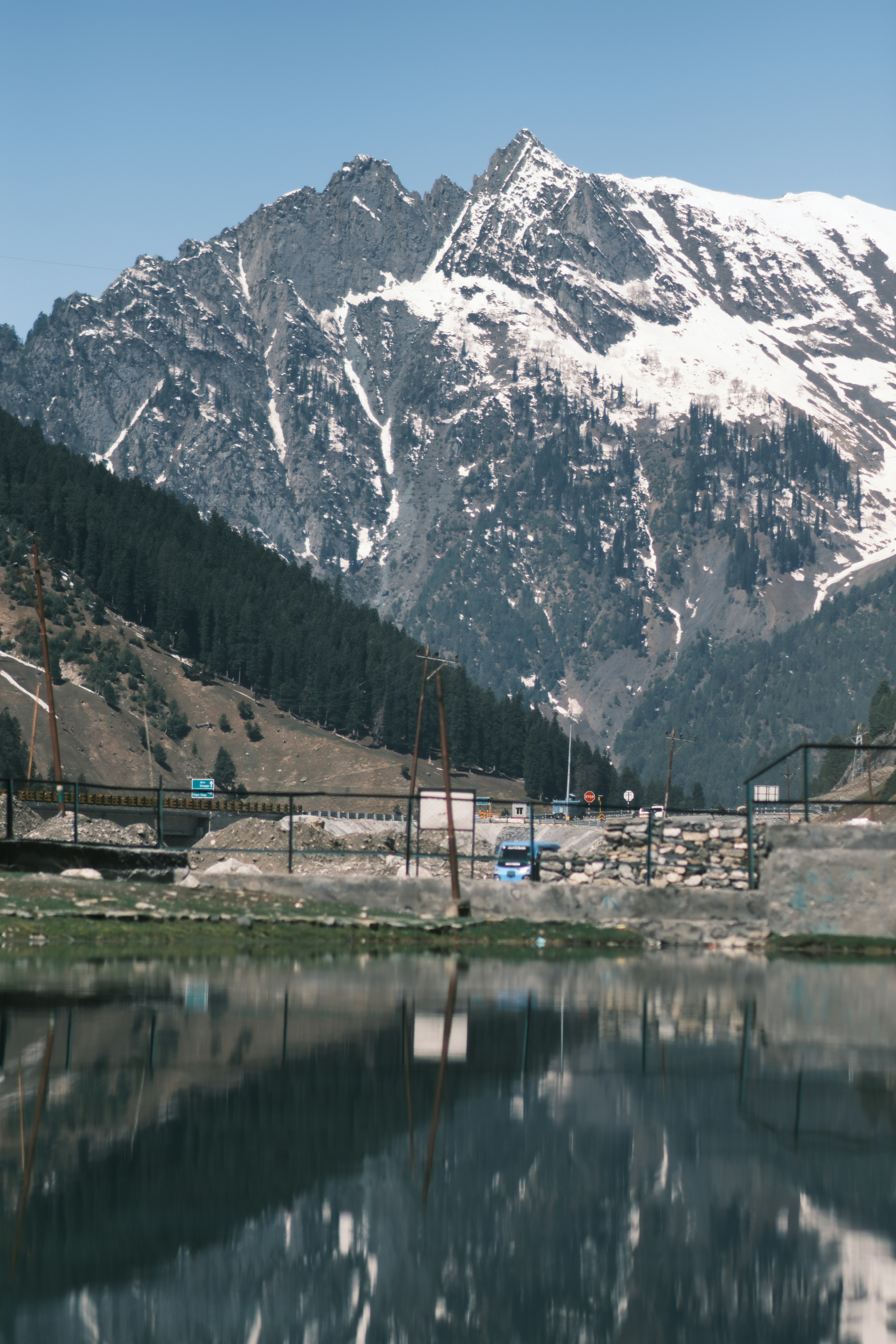 Snow-capped mountains reflecting in a calm pond with a road and forest nearby