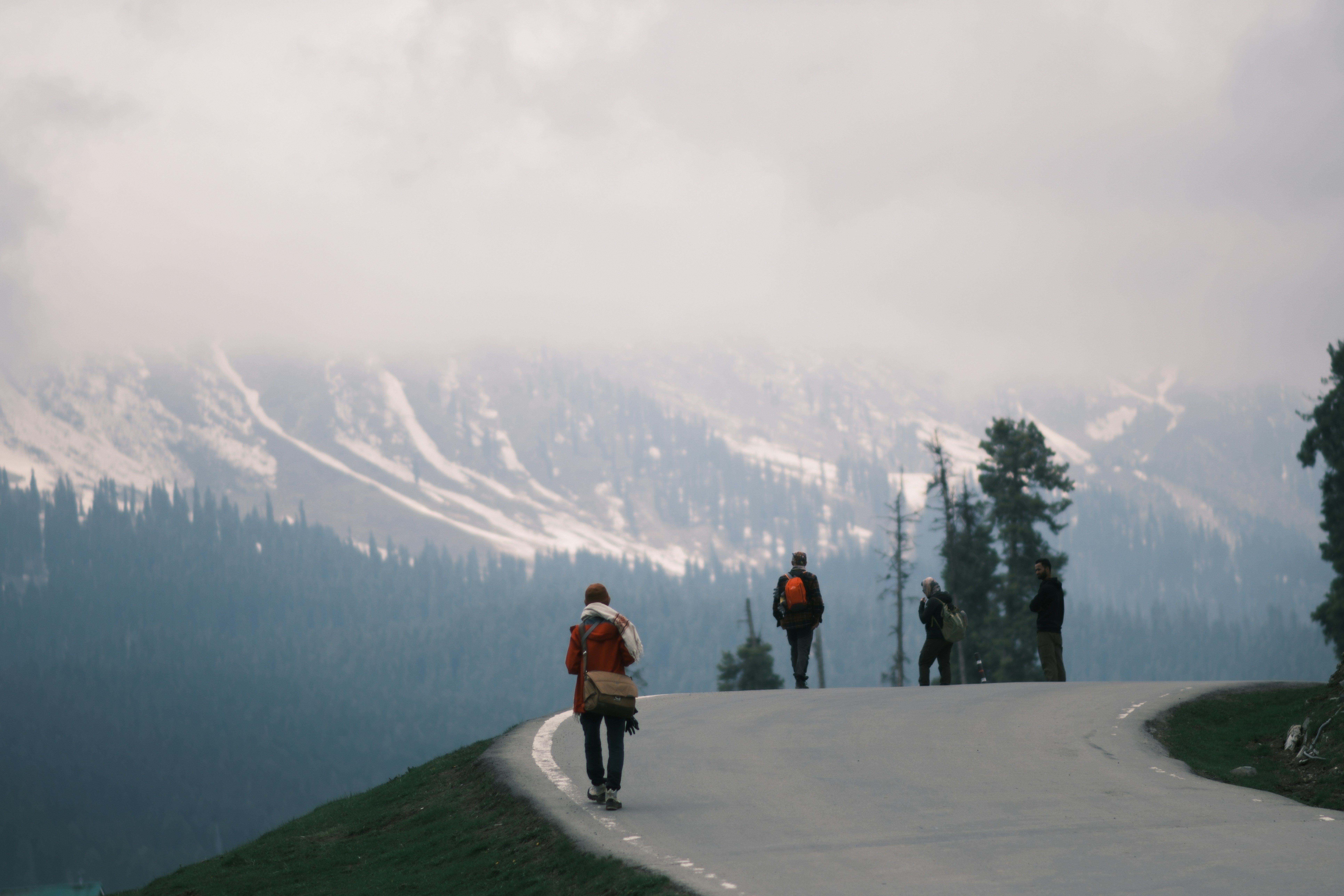 Four people walking along a mountain road with snow-capped peaks in the distance