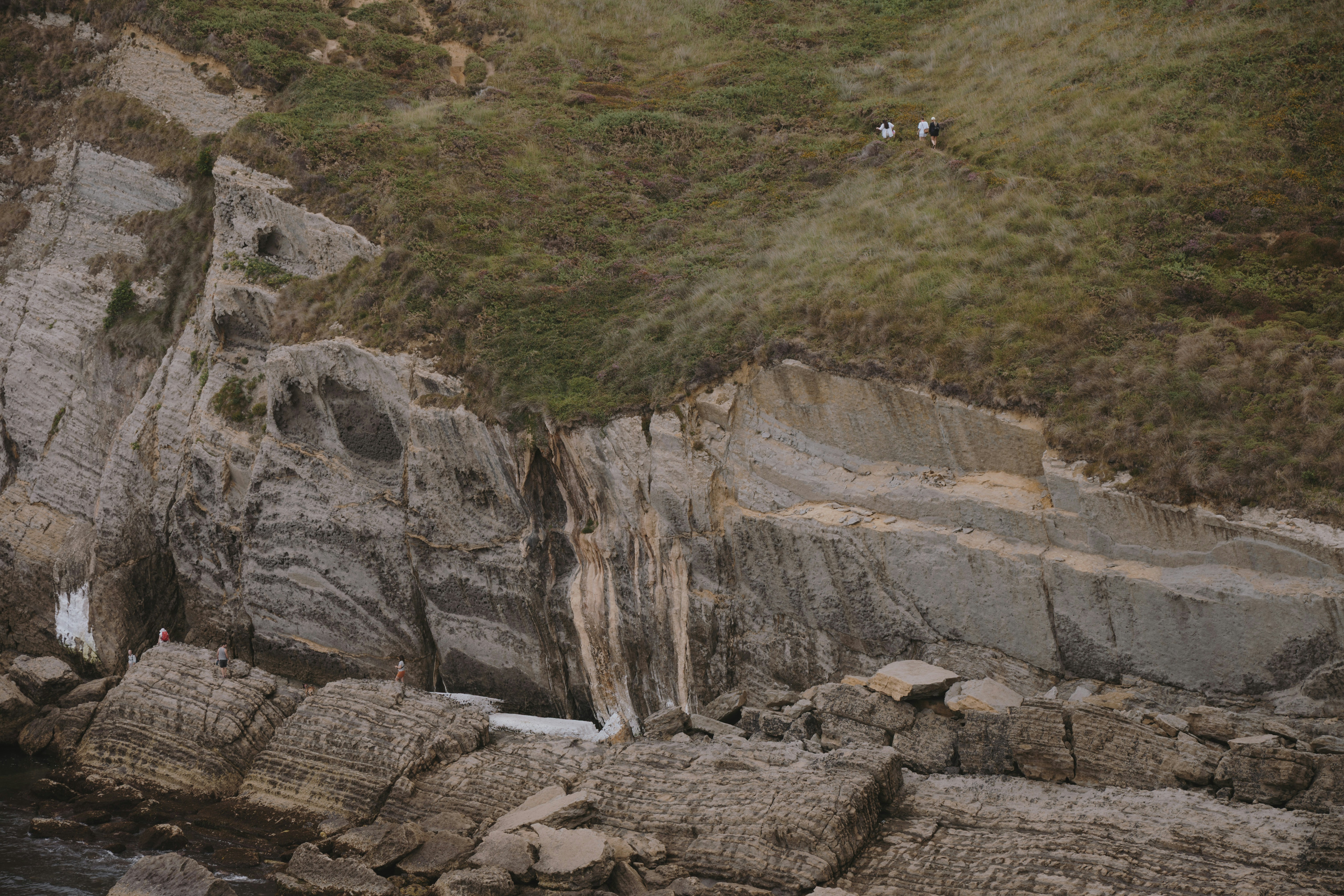 People walking on a grassy cliff above layered sedimentary rock formations