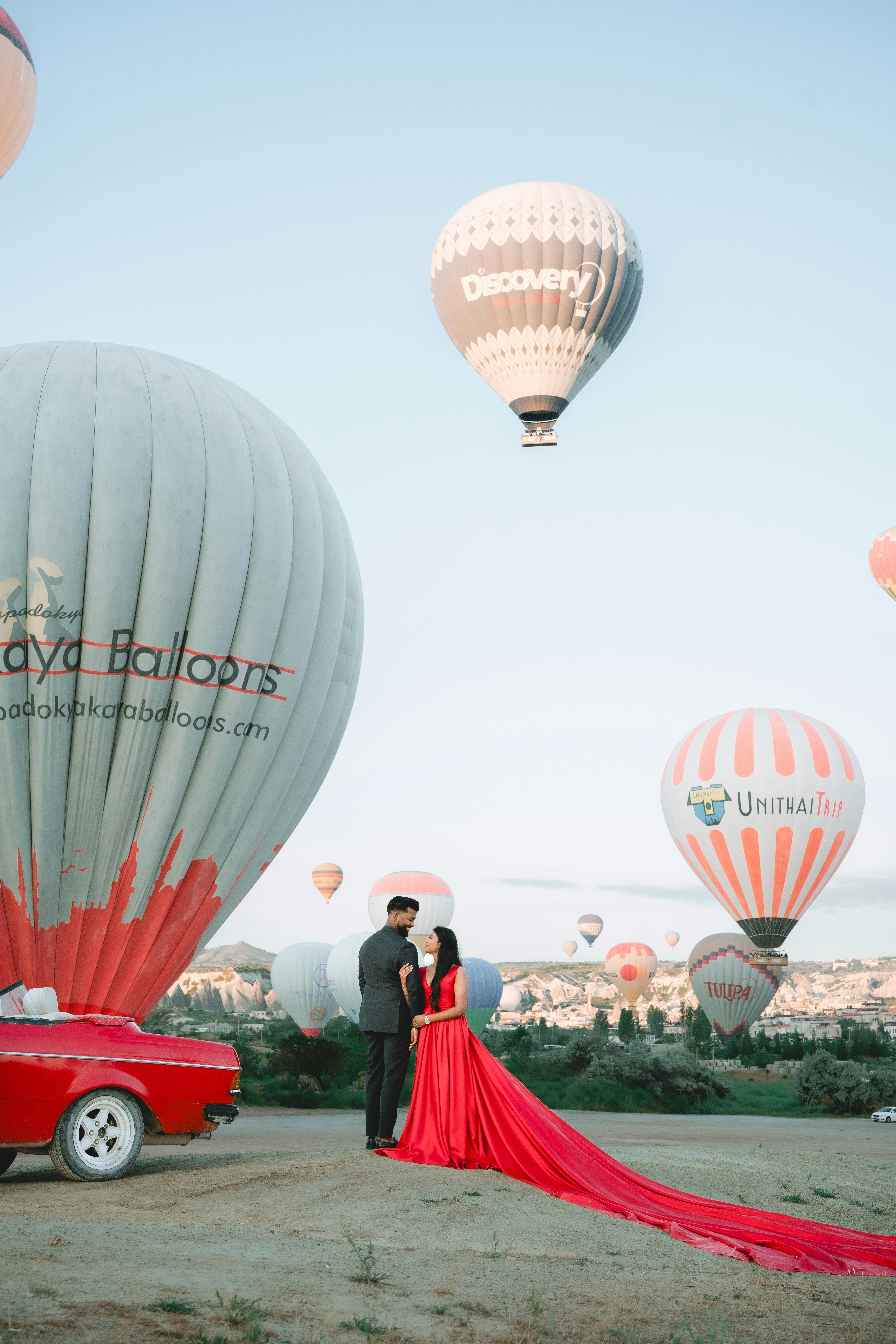 A couple in formal attire standing near a red vintage car with hot air balloons