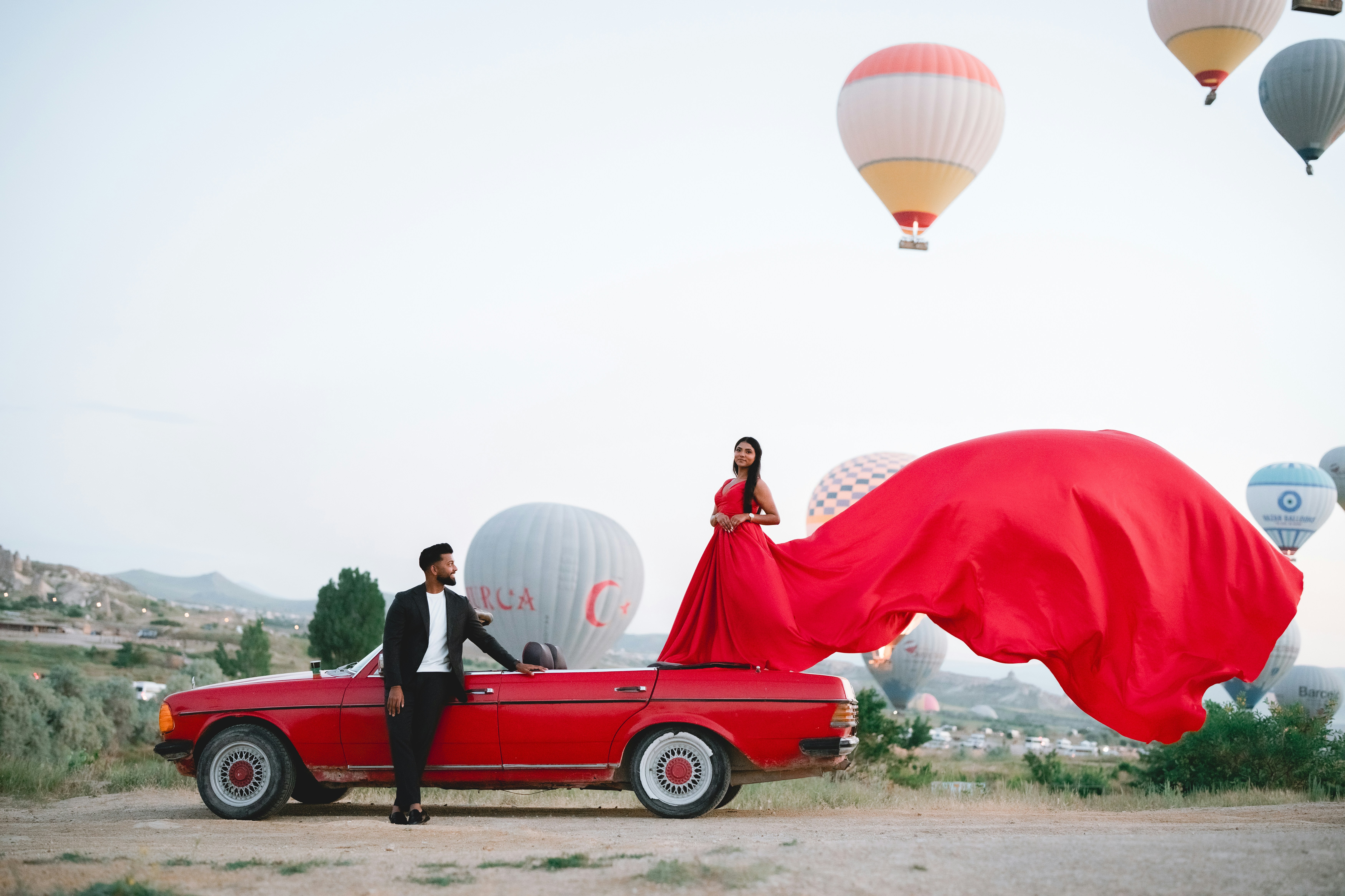A woman in a red dress on a red convertible under hot air balloons