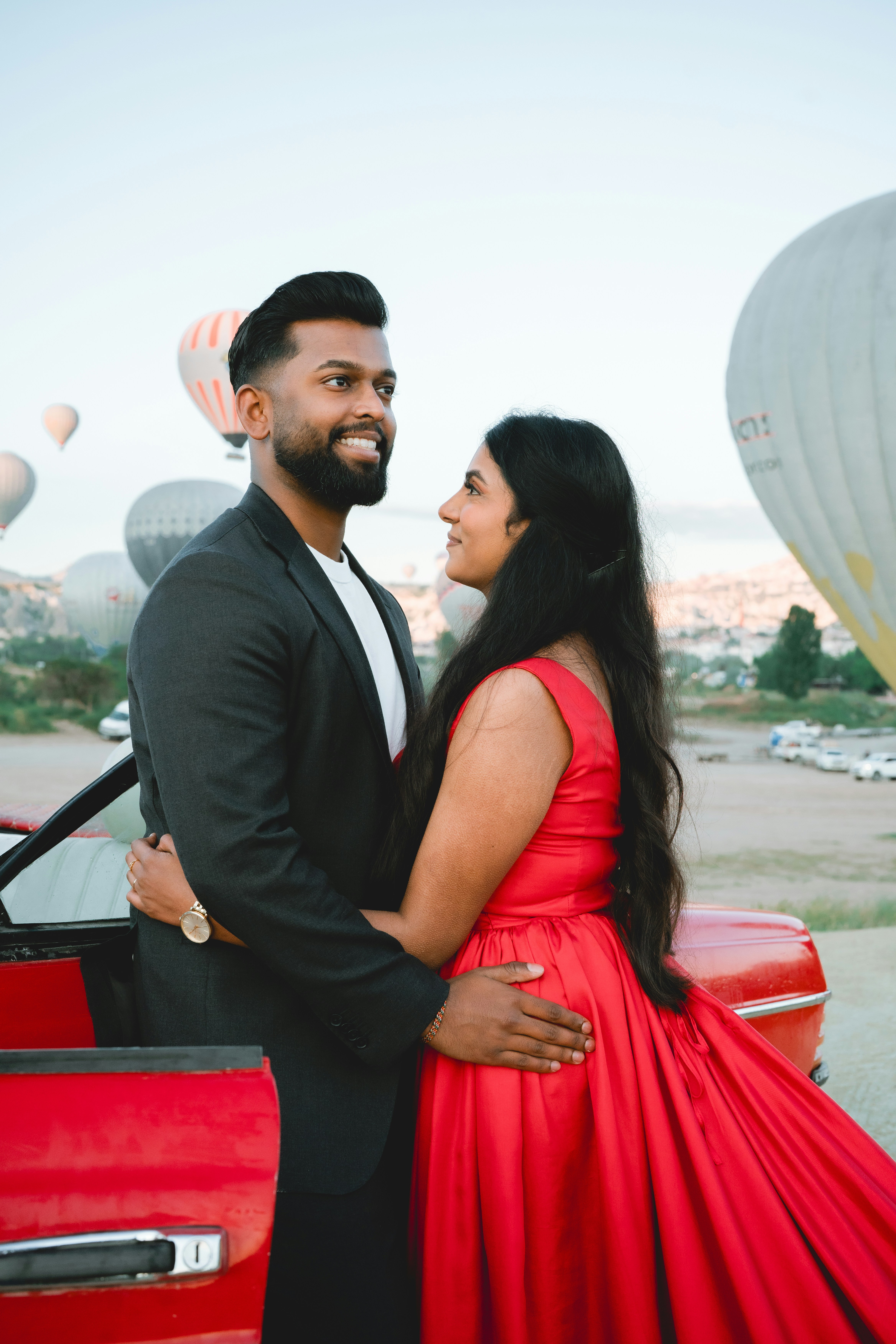 A man and a woman embracing beside a red convertible with hot air balloons
