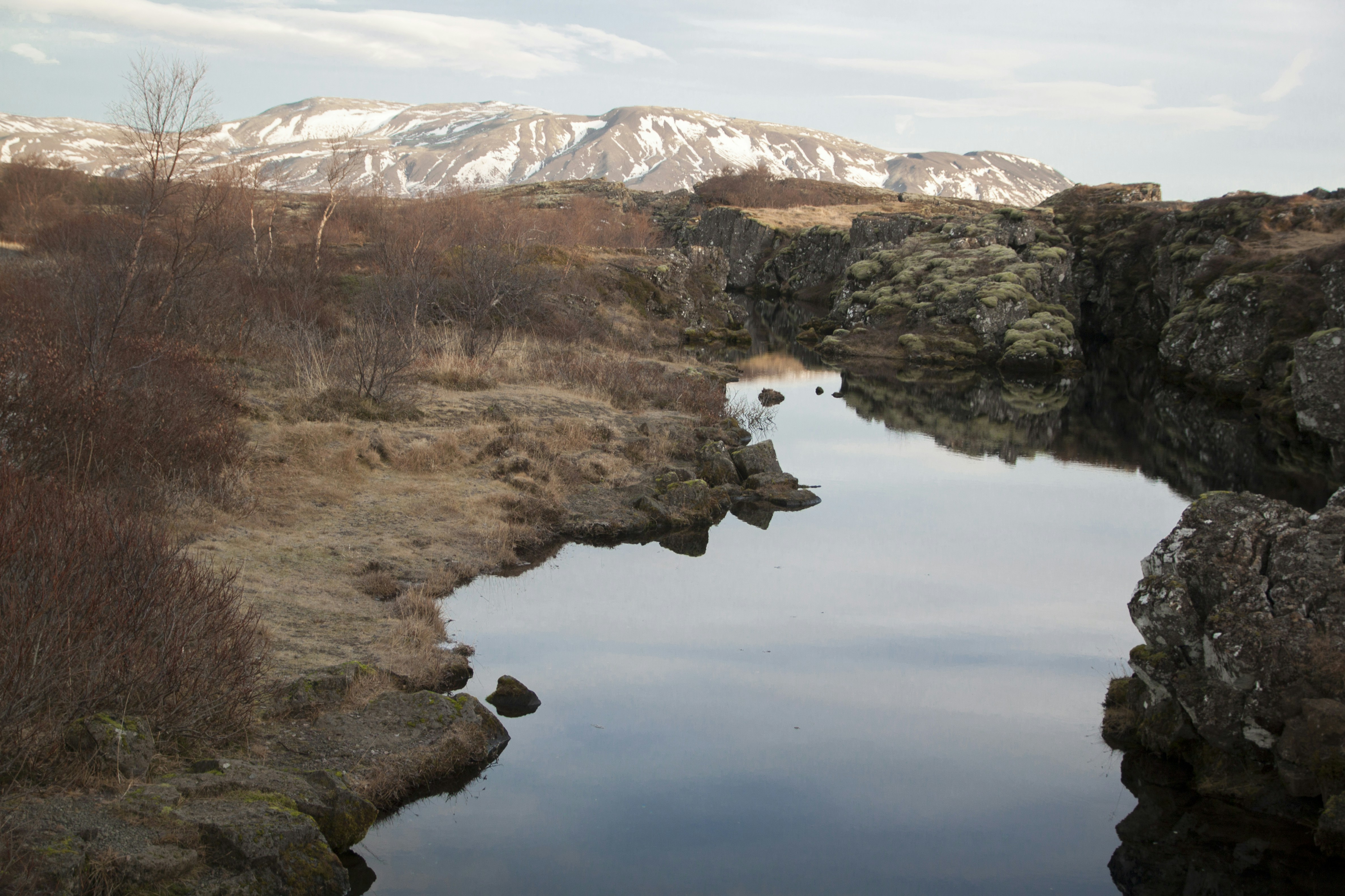 A calm river flowing through a rocky mossy gorge with snow-capped mountains