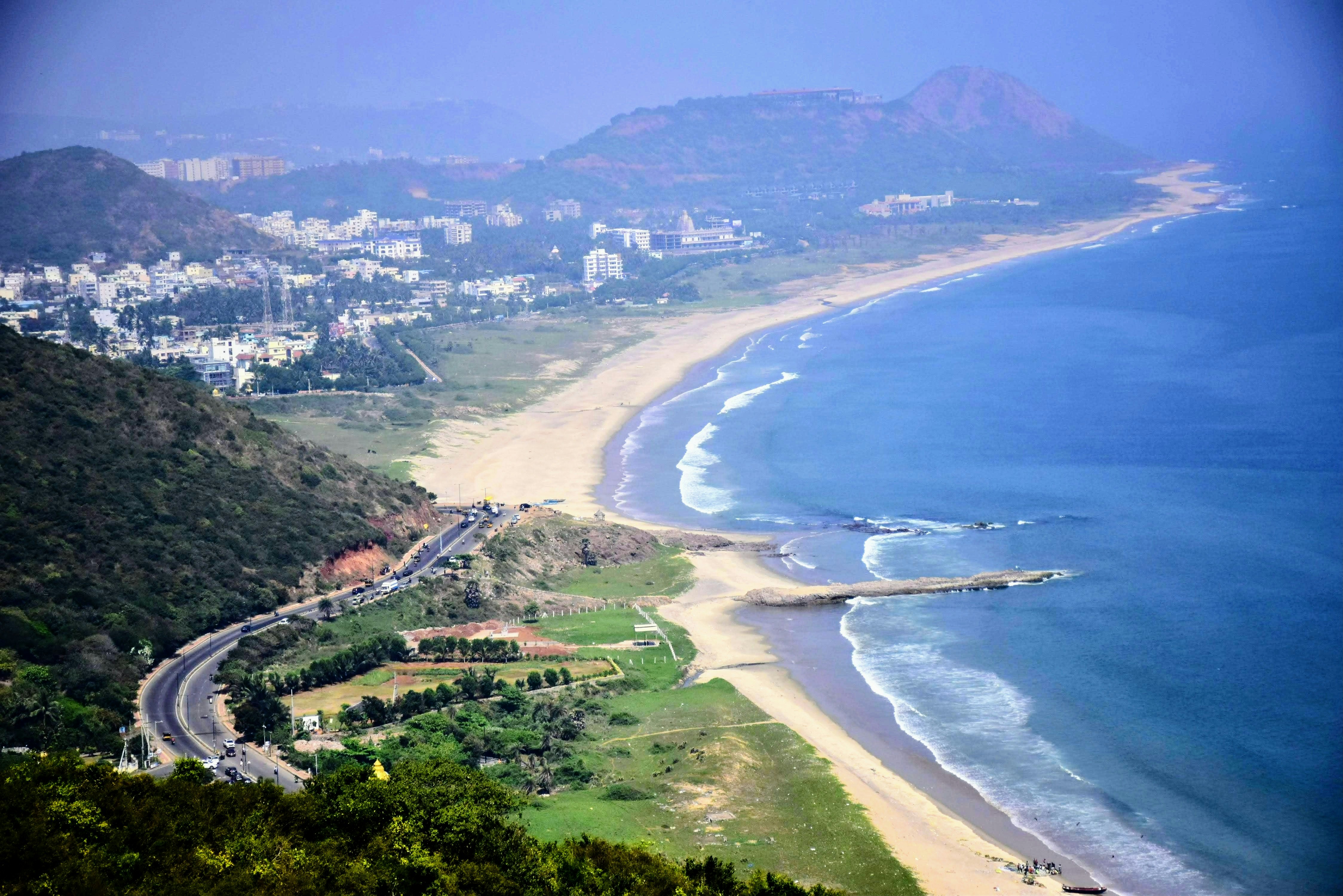 A coastal road winding along a sandy beach below green hills in Visakhapatnam