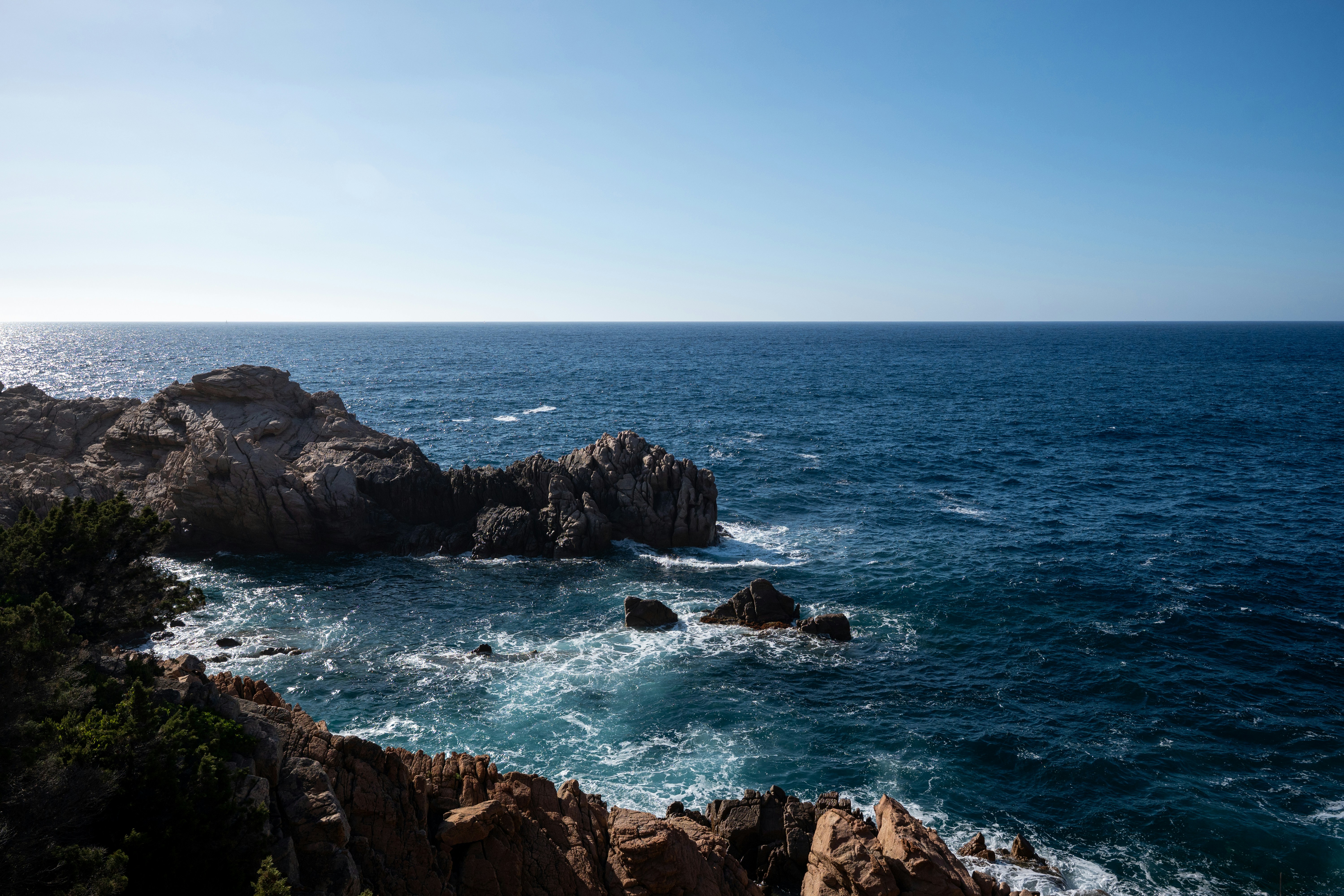 Waves crashing against jagged brown rocks along a deep blue sea coastline