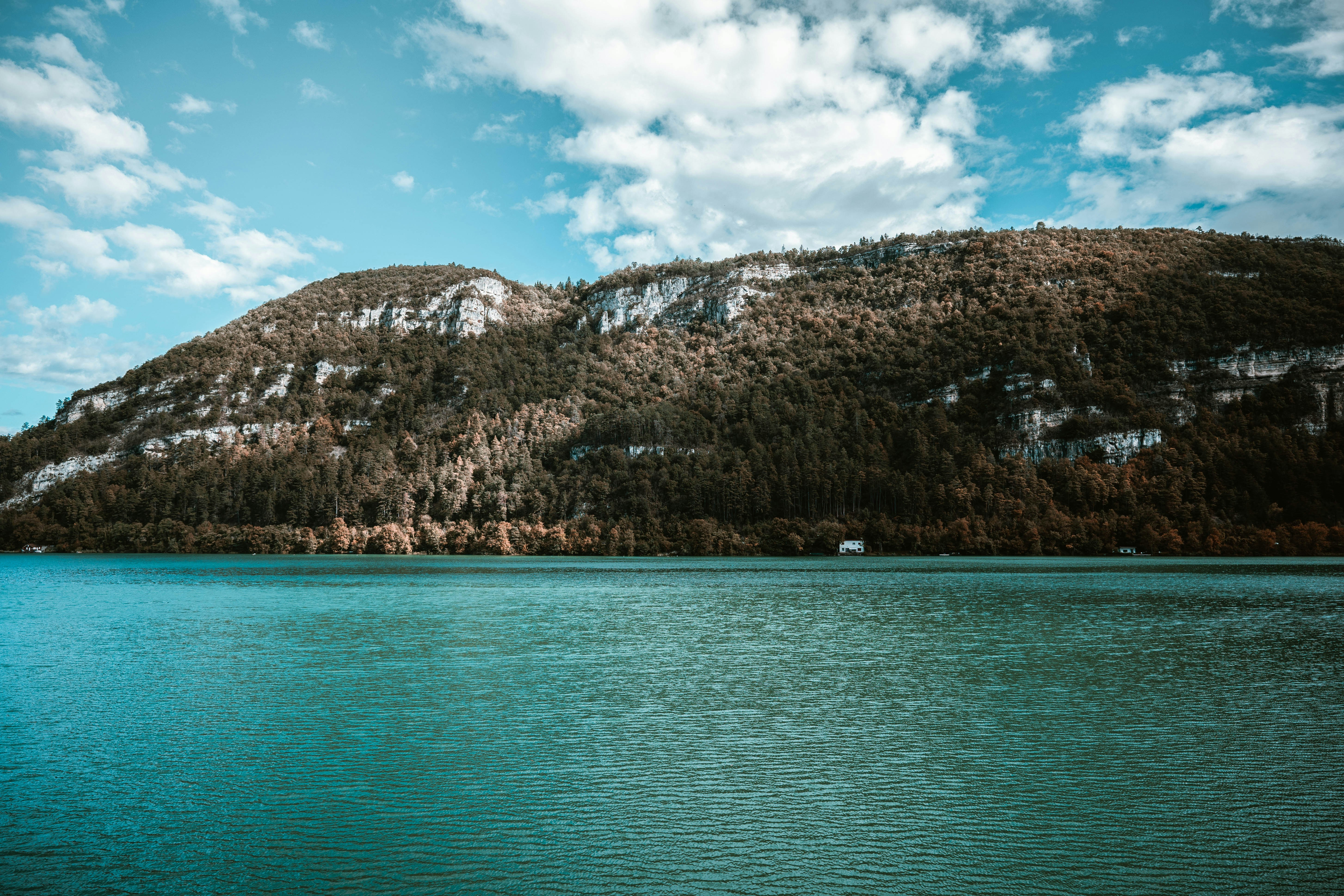 A turquoise lake beneath a forested mountain with rocky cliffs under a blue sky