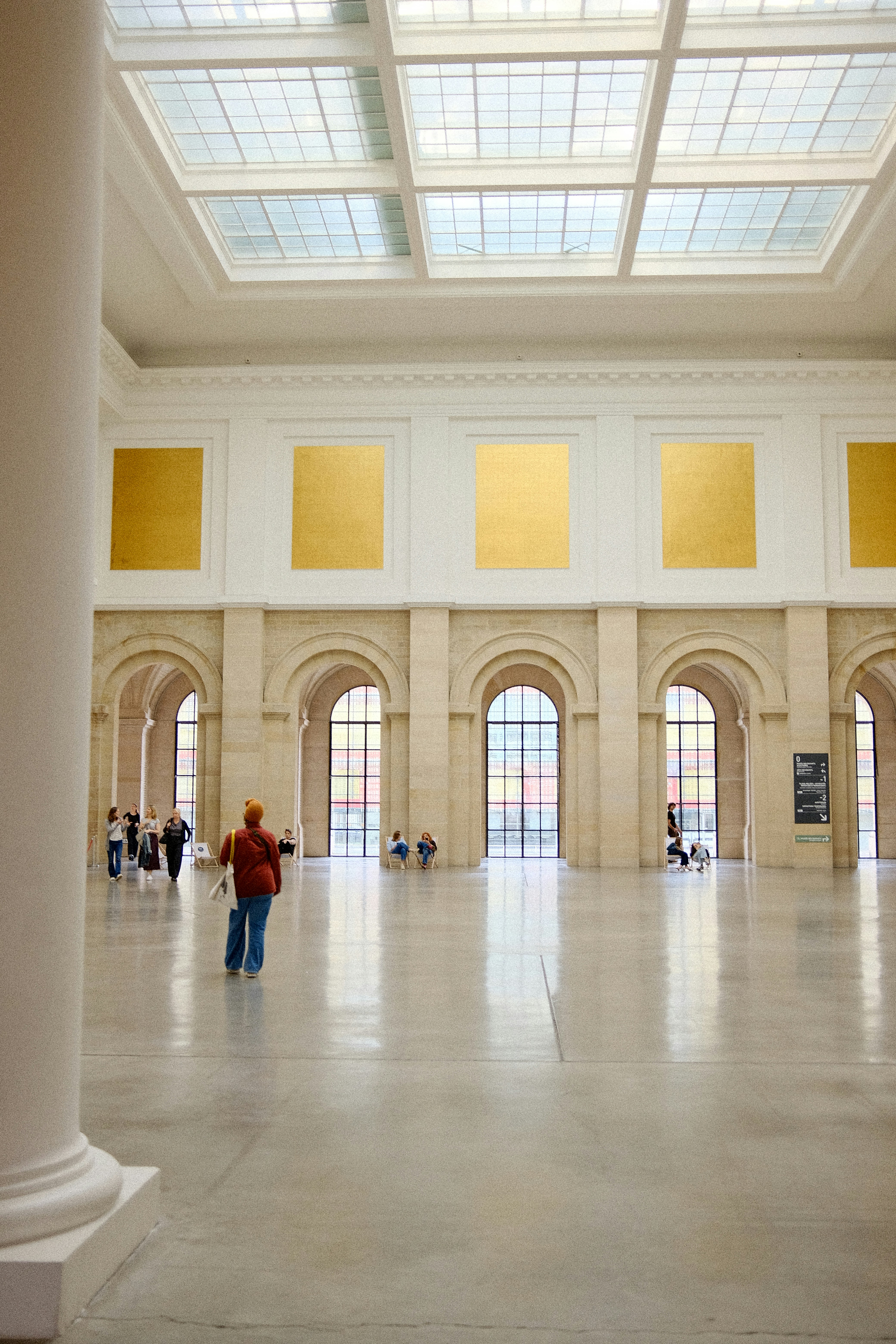 A person in a red jacket walking through a spacious museum hall with arched windows