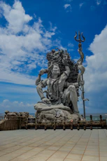 A large grey stone statue of Shiva holding a trident against a blue sky