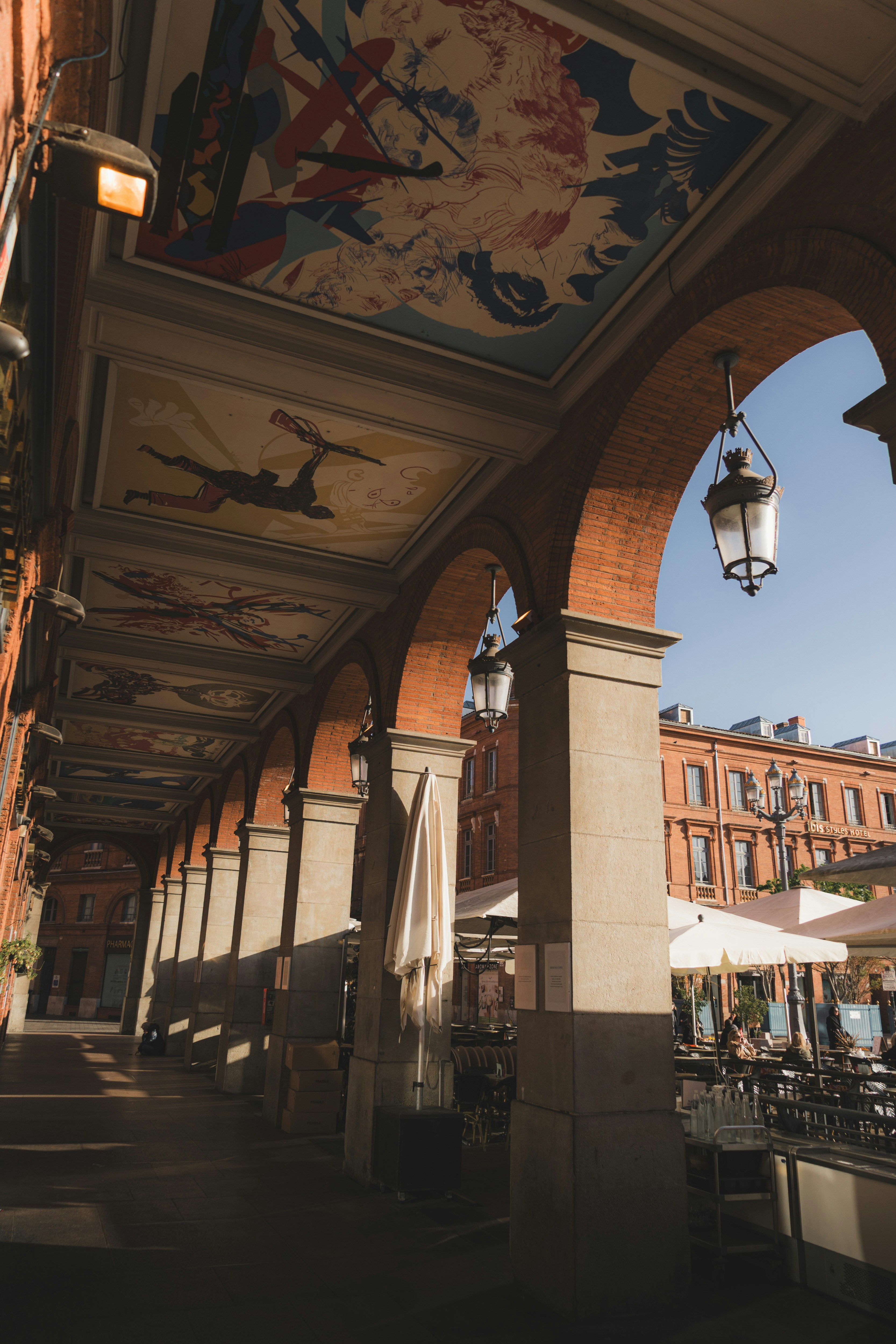 Brick arches with painted ceilings and hanging lanterns overlooking a sunlit outdoor cafe