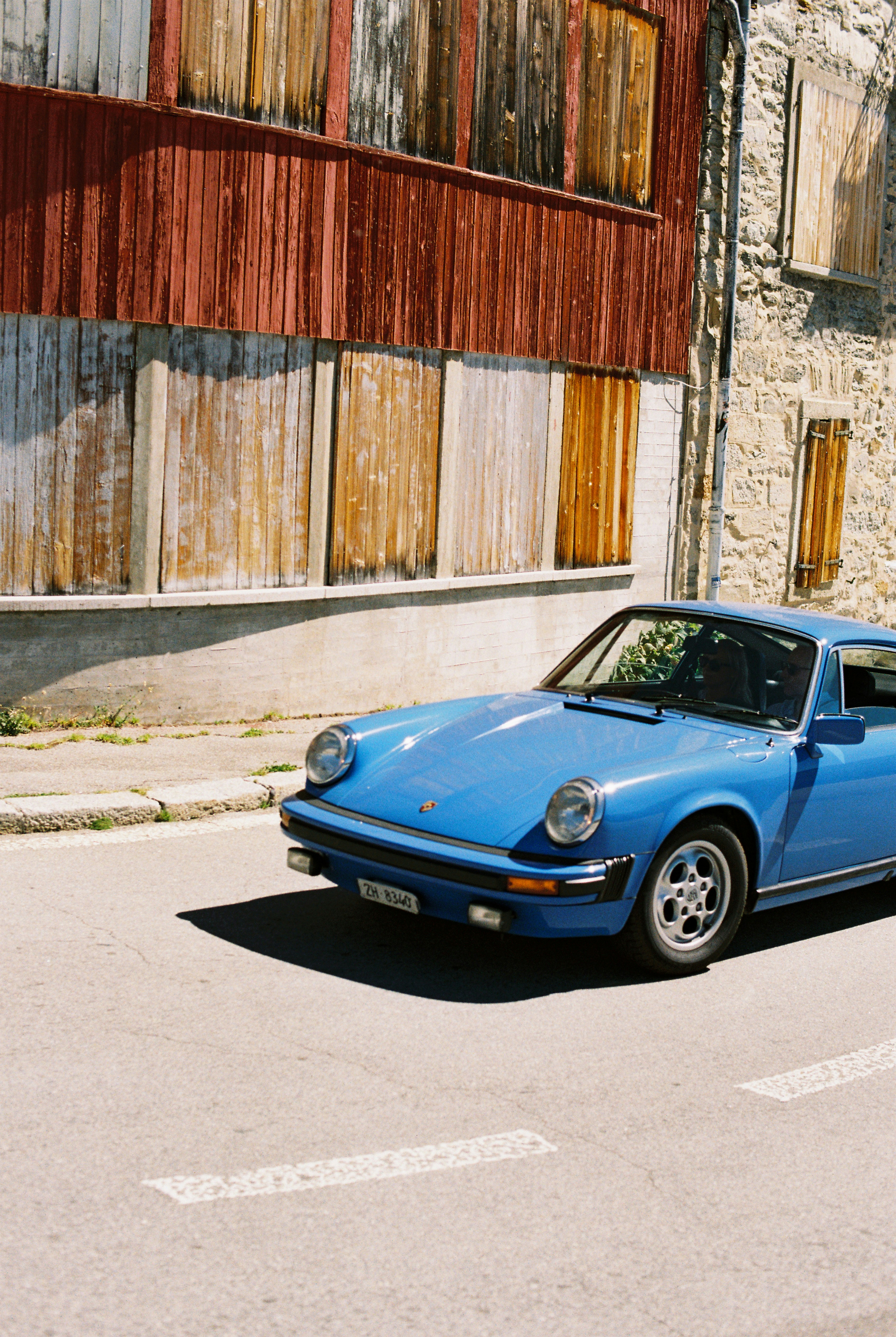 A blue vintage sports car driving past a stone building with wooden shutters