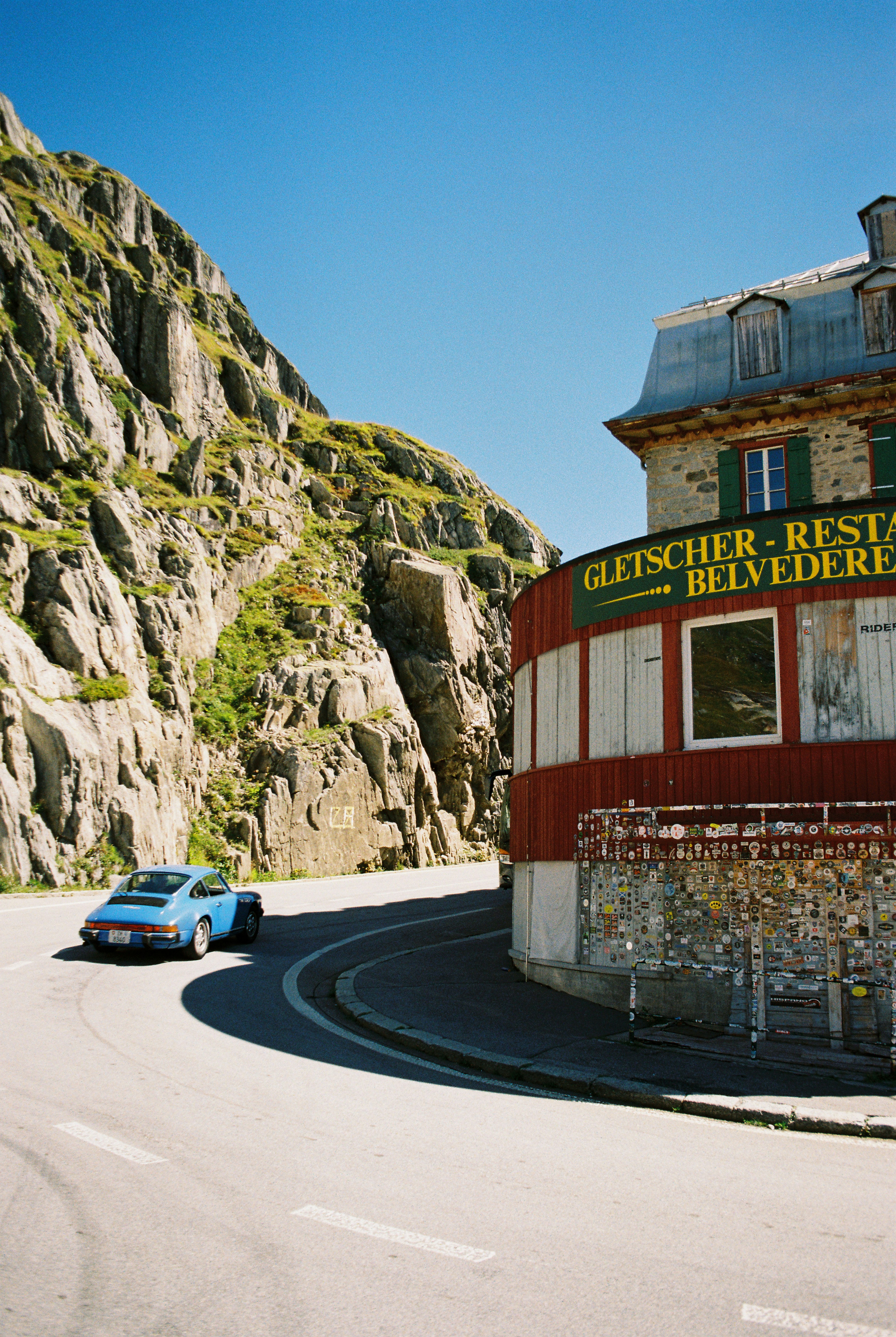 A blue vintage sports car driving past the Hotel Belvedere on a mountain road