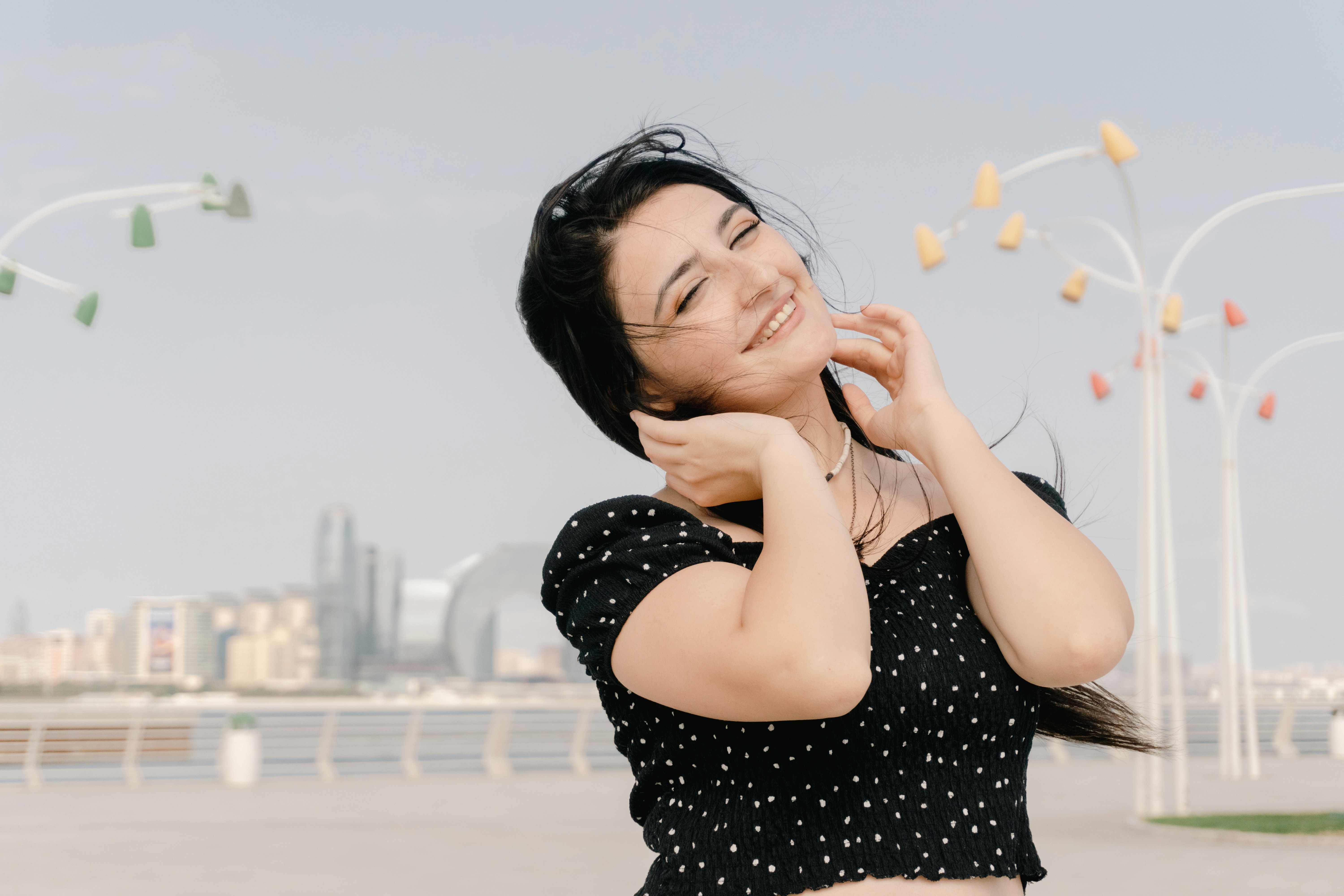 A woman in a black polka dot top touching her hair on a waterfront promenade