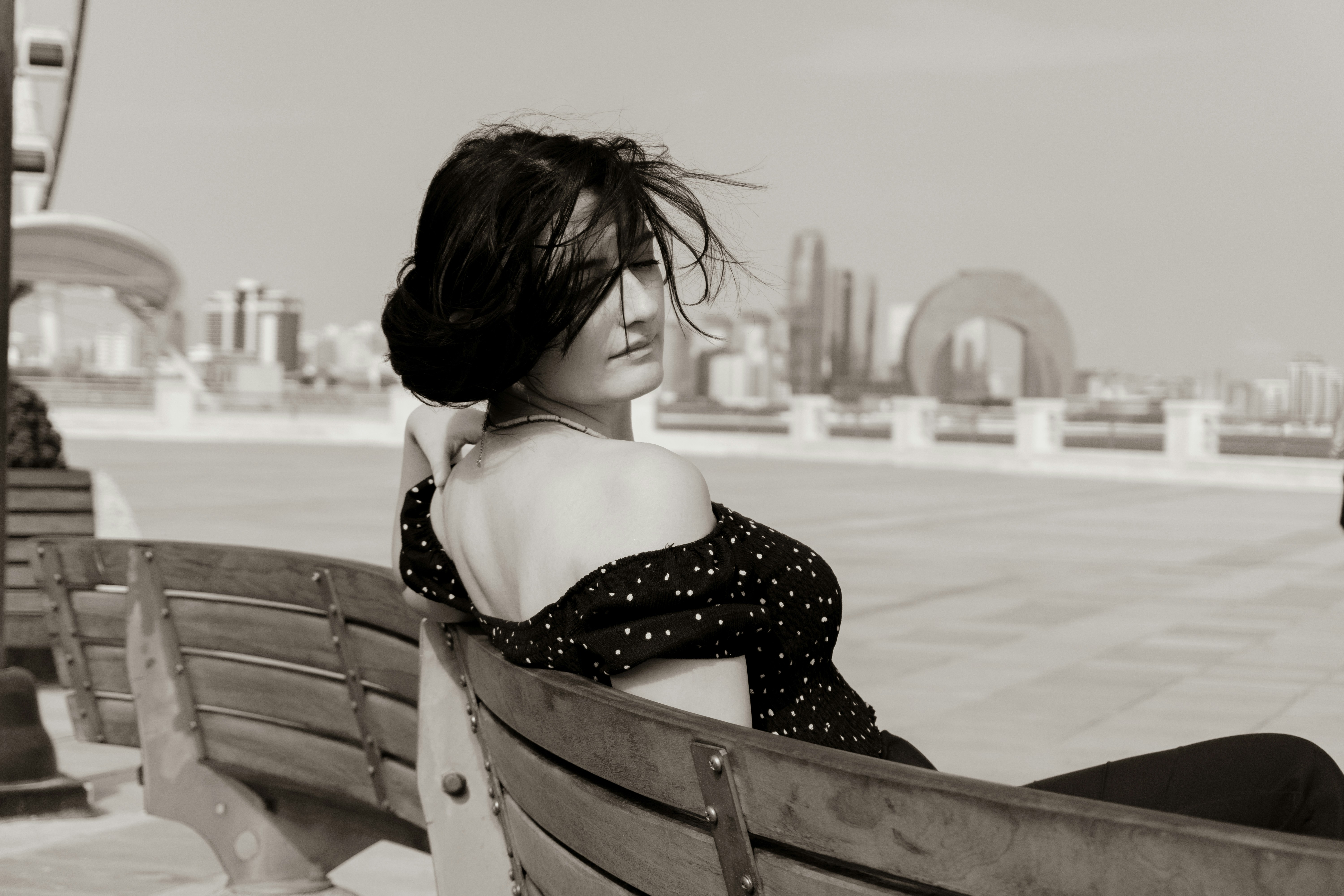A woman in a polka dot top on a wooden bench with city buildings