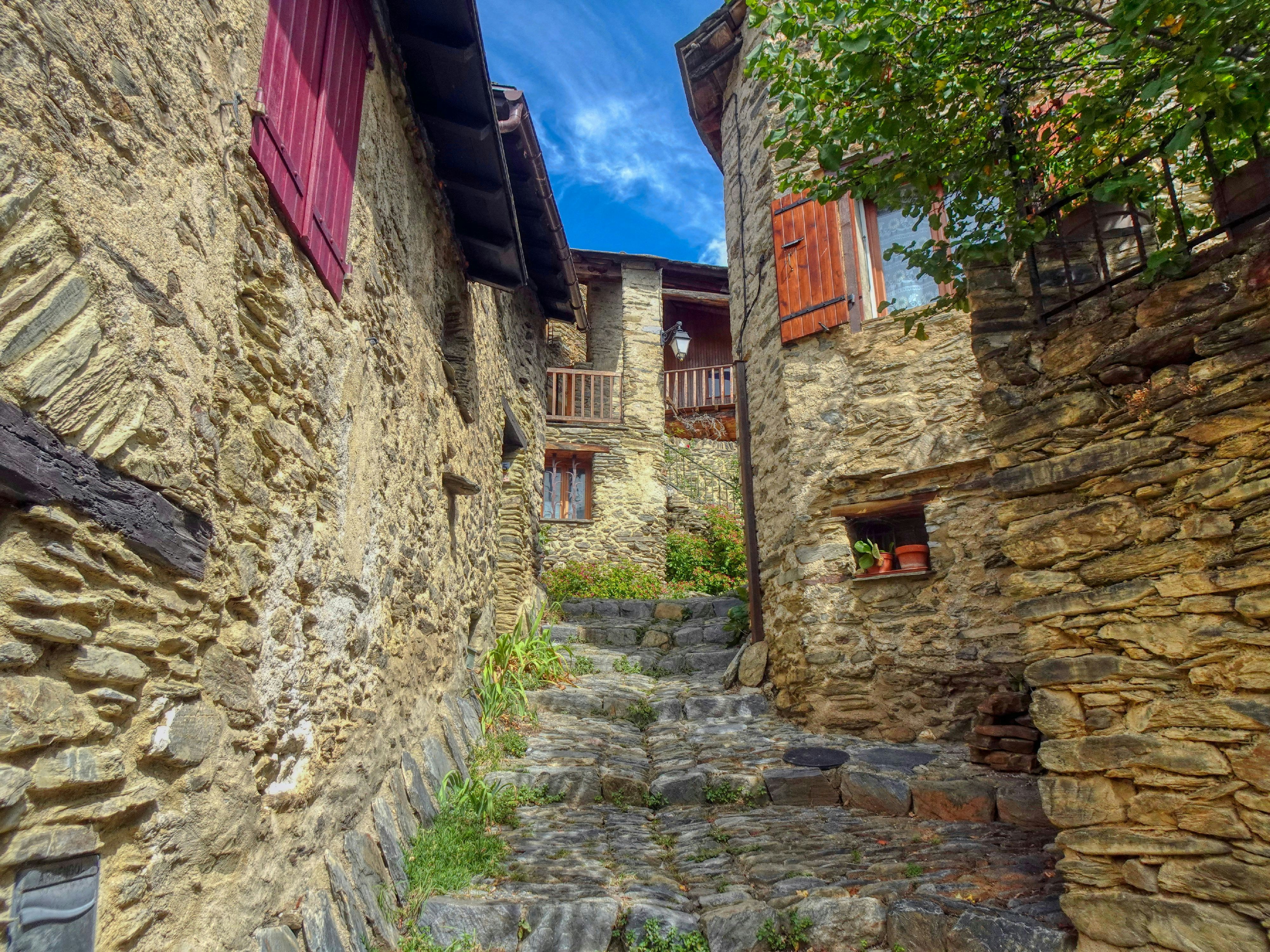 Eine schmale Steingasse mit rustikalen Gebäuden und roten Fensterläden unter blauem Himmel