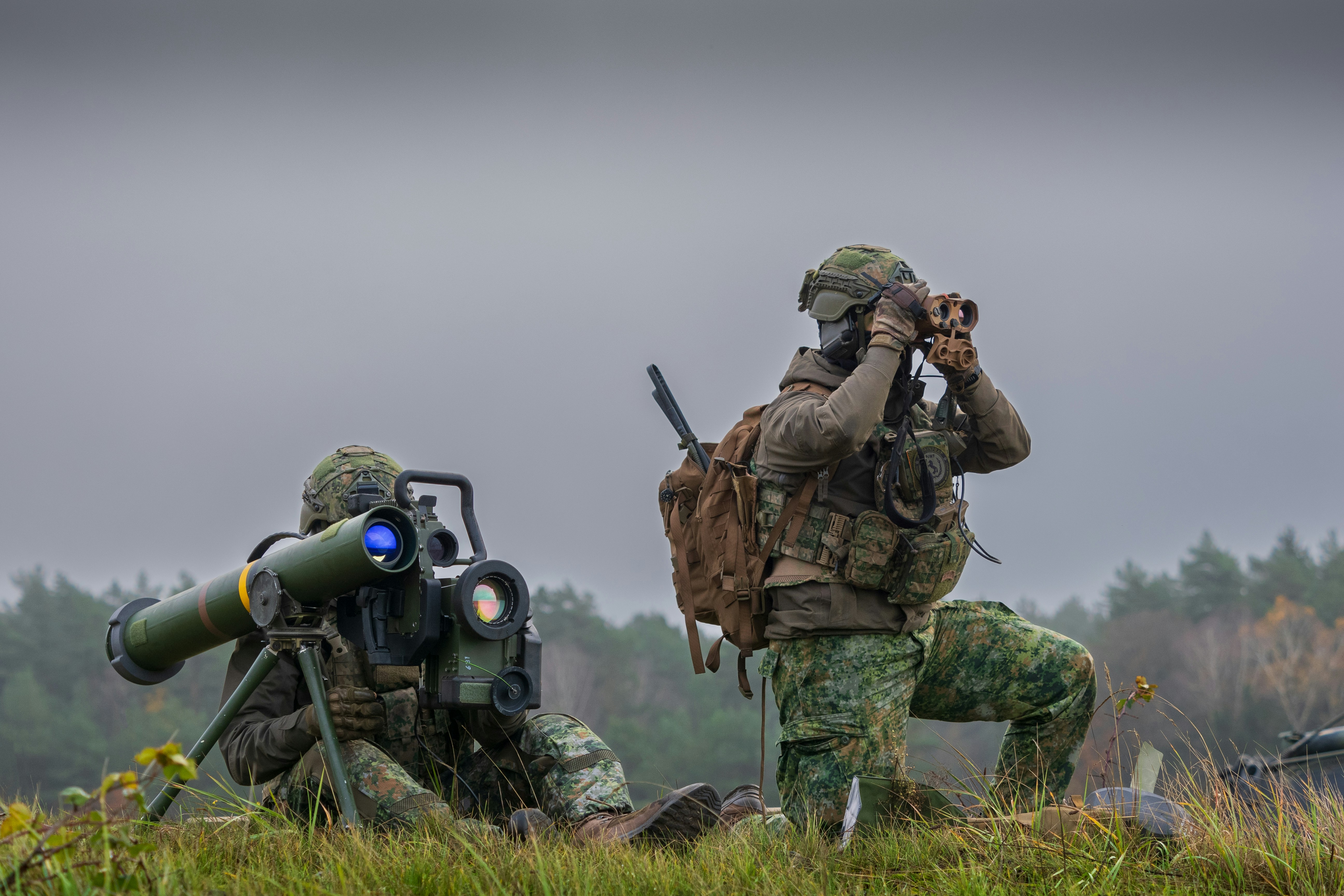 Two soldiers in camouflage gear with a missile launcher and binoculars in a field