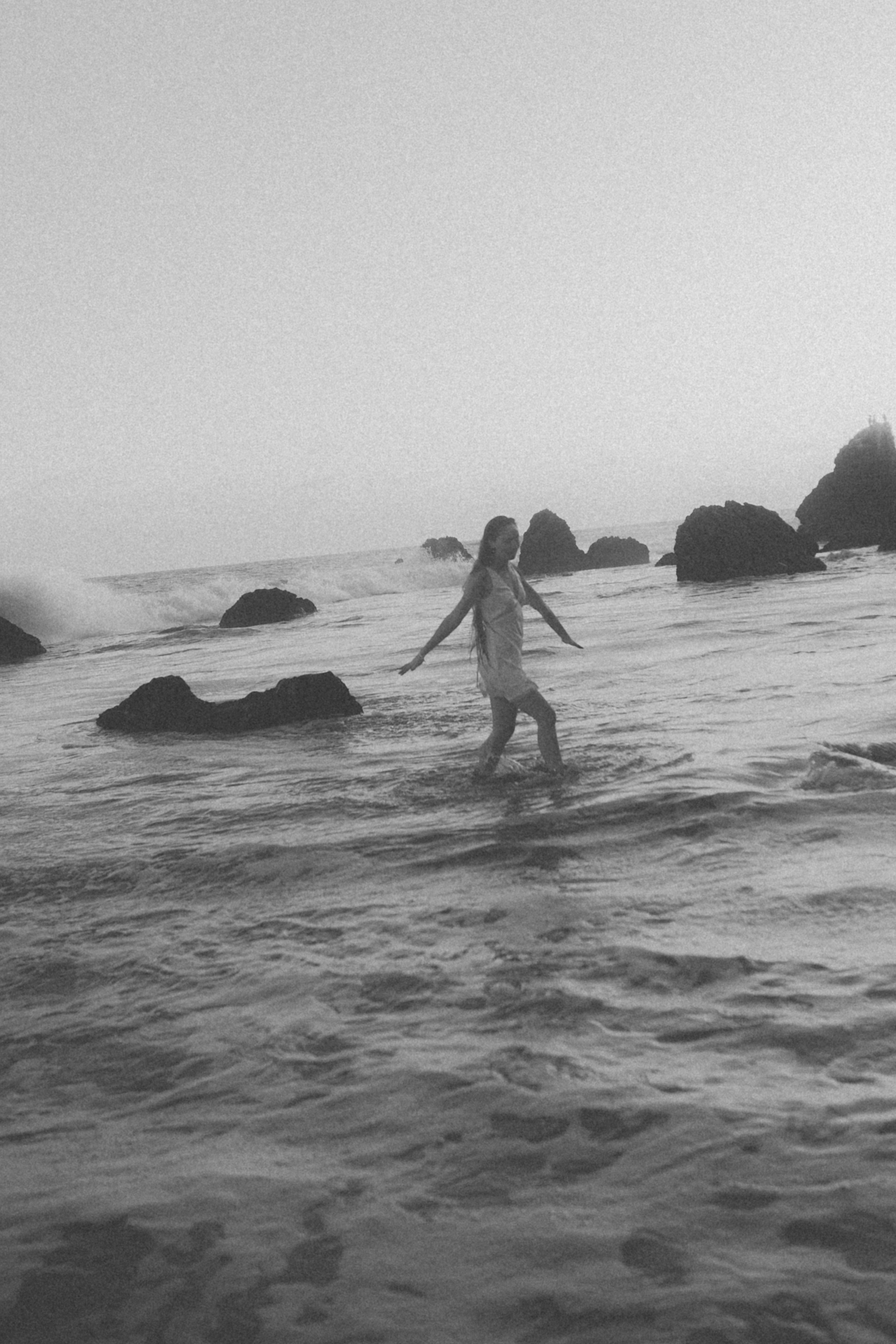A woman in a light dress wading through ocean water near rocky outcrops