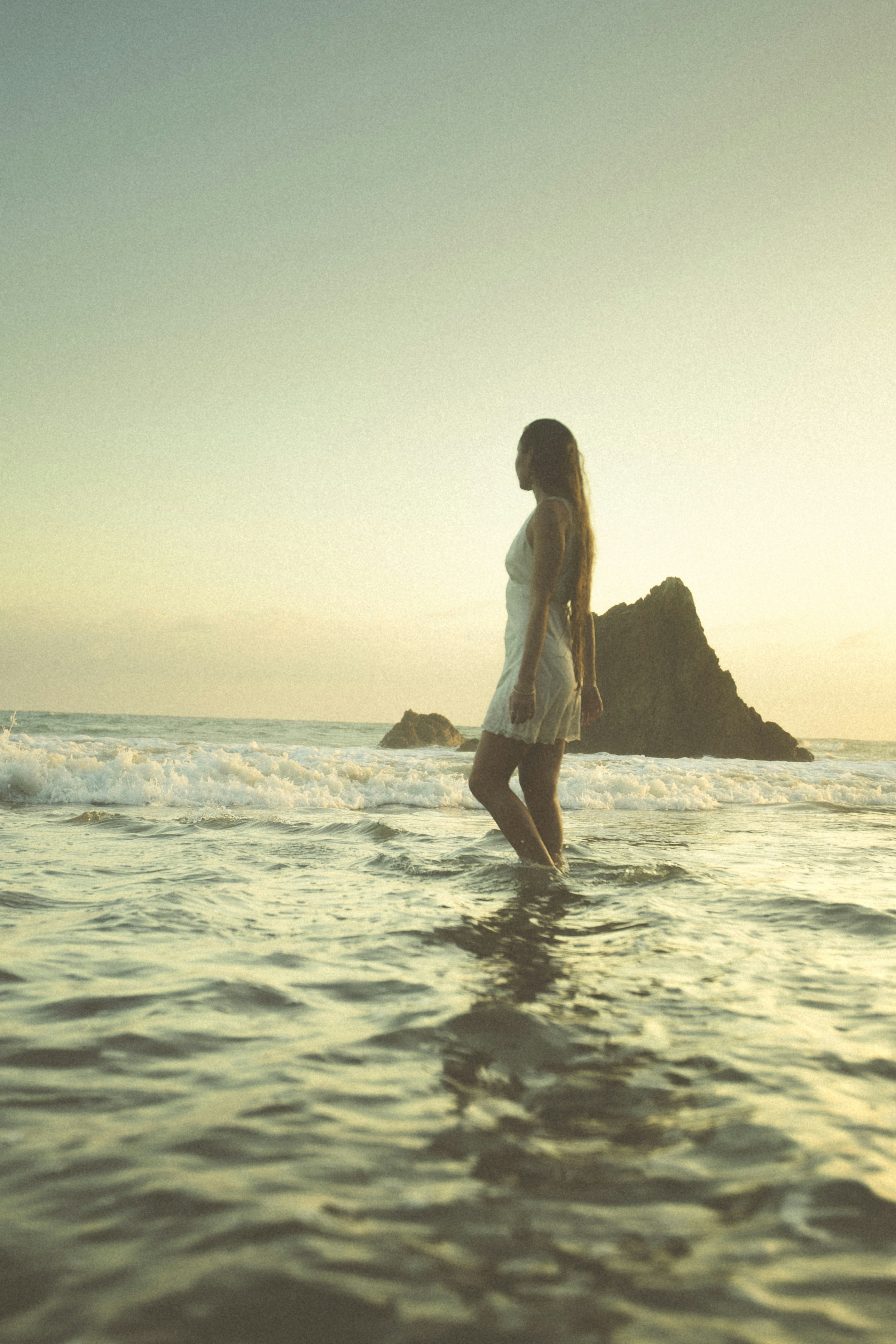 A woman in a white dress walking through shallow ocean waves at golden hour