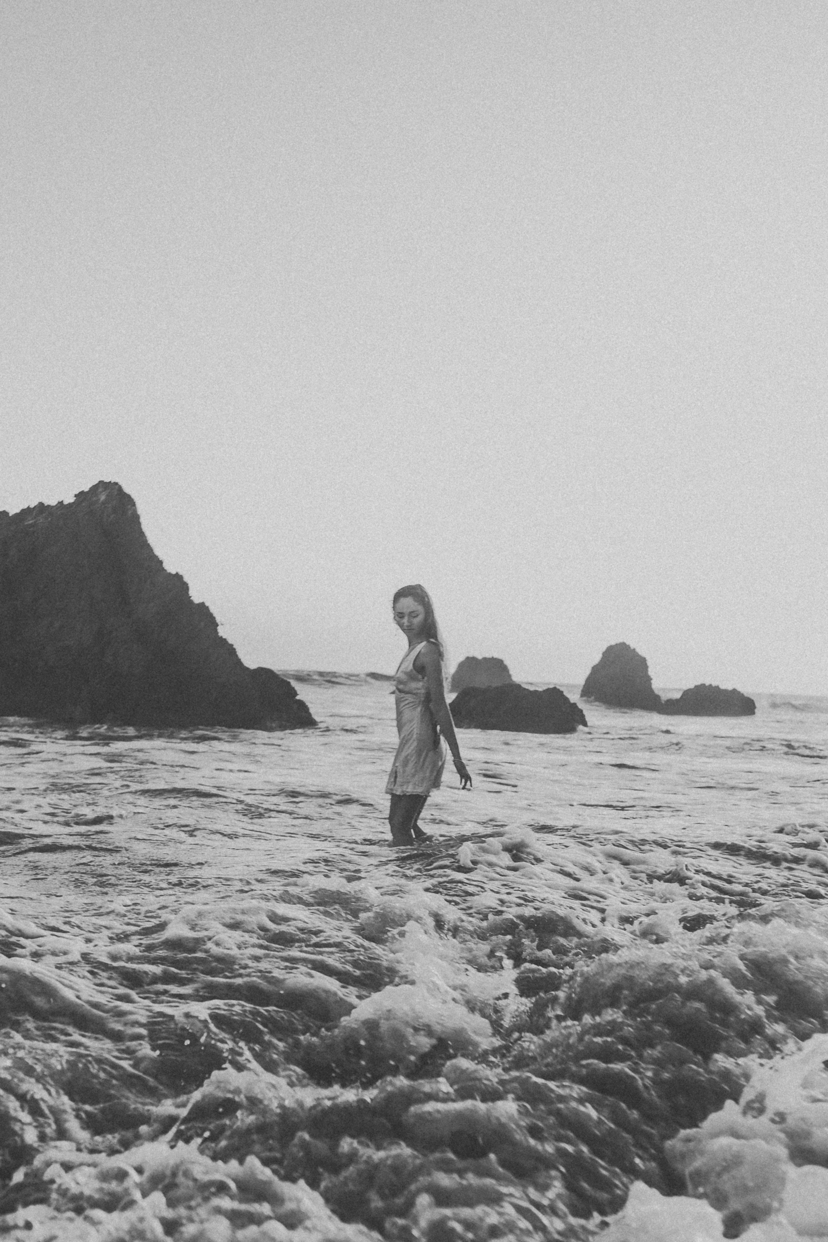 A woman in a light dress wading through ocean waves near rocky outcrops