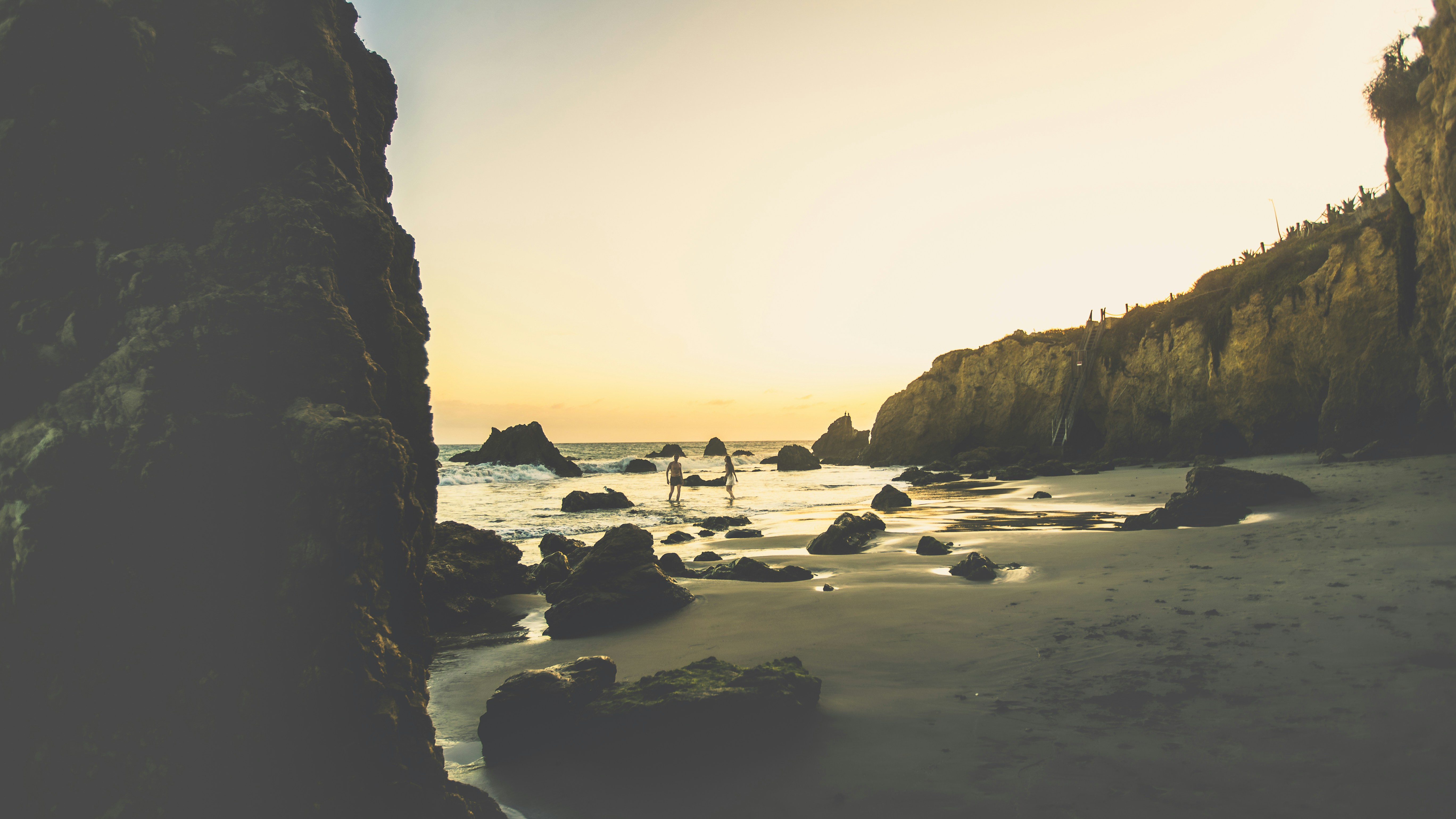 Two people wading in the ocean near rocky cliffs under a warm low sun