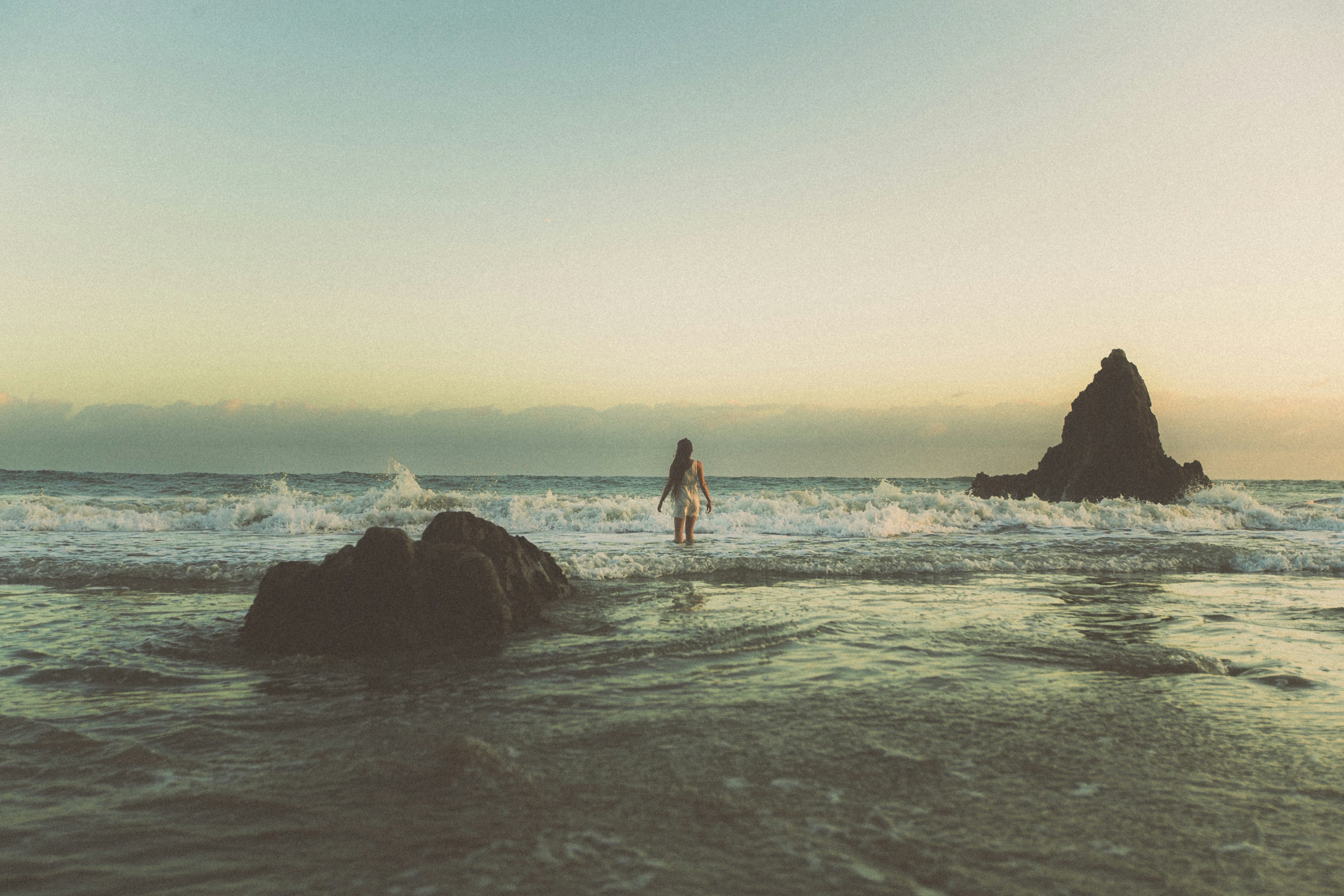A woman in a white dress wading into ocean waves near rocky outcrops