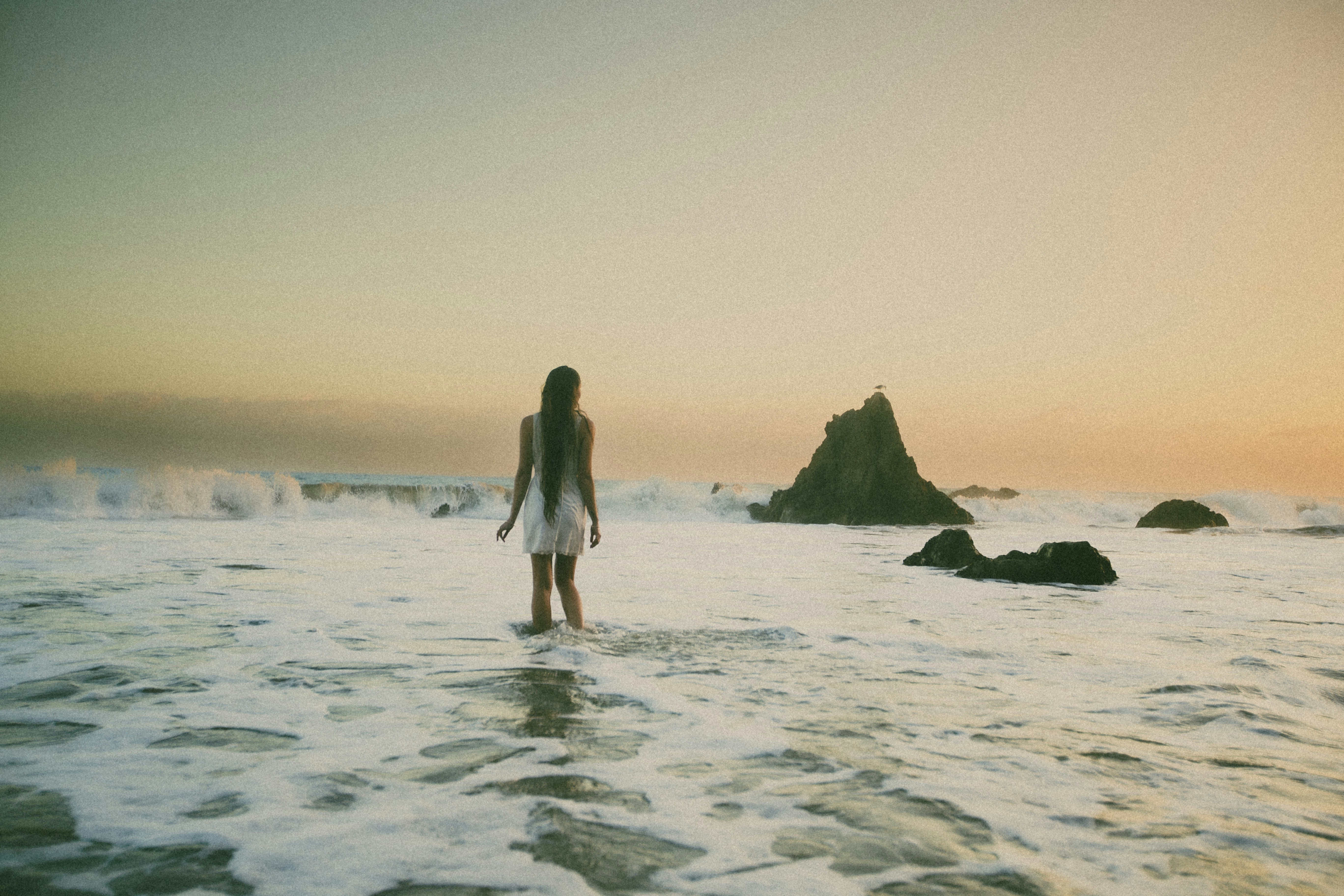 A woman in a white dress wading through ocean surf at golden hour