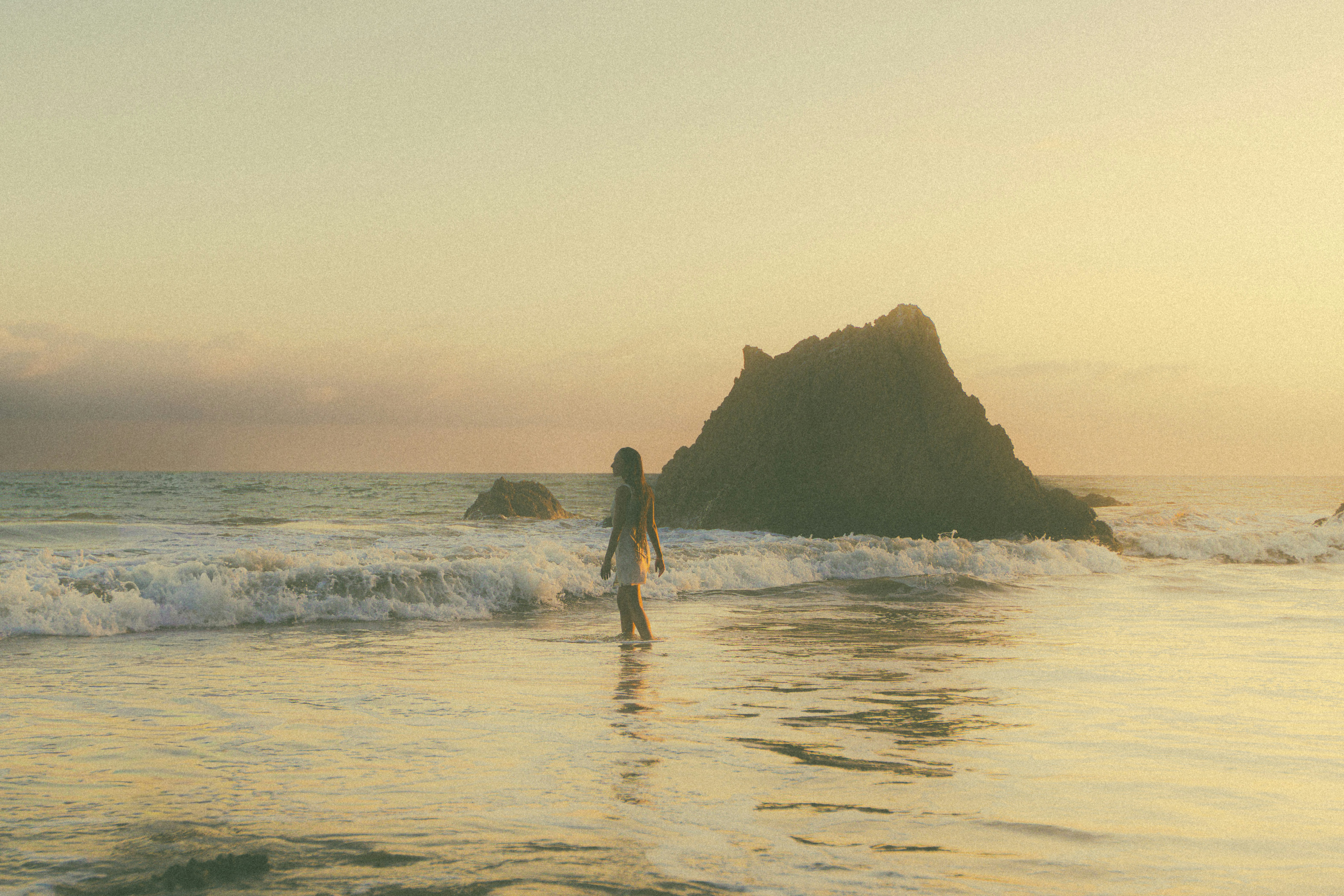 A person walking in shallow ocean water near a large rock at golden hour