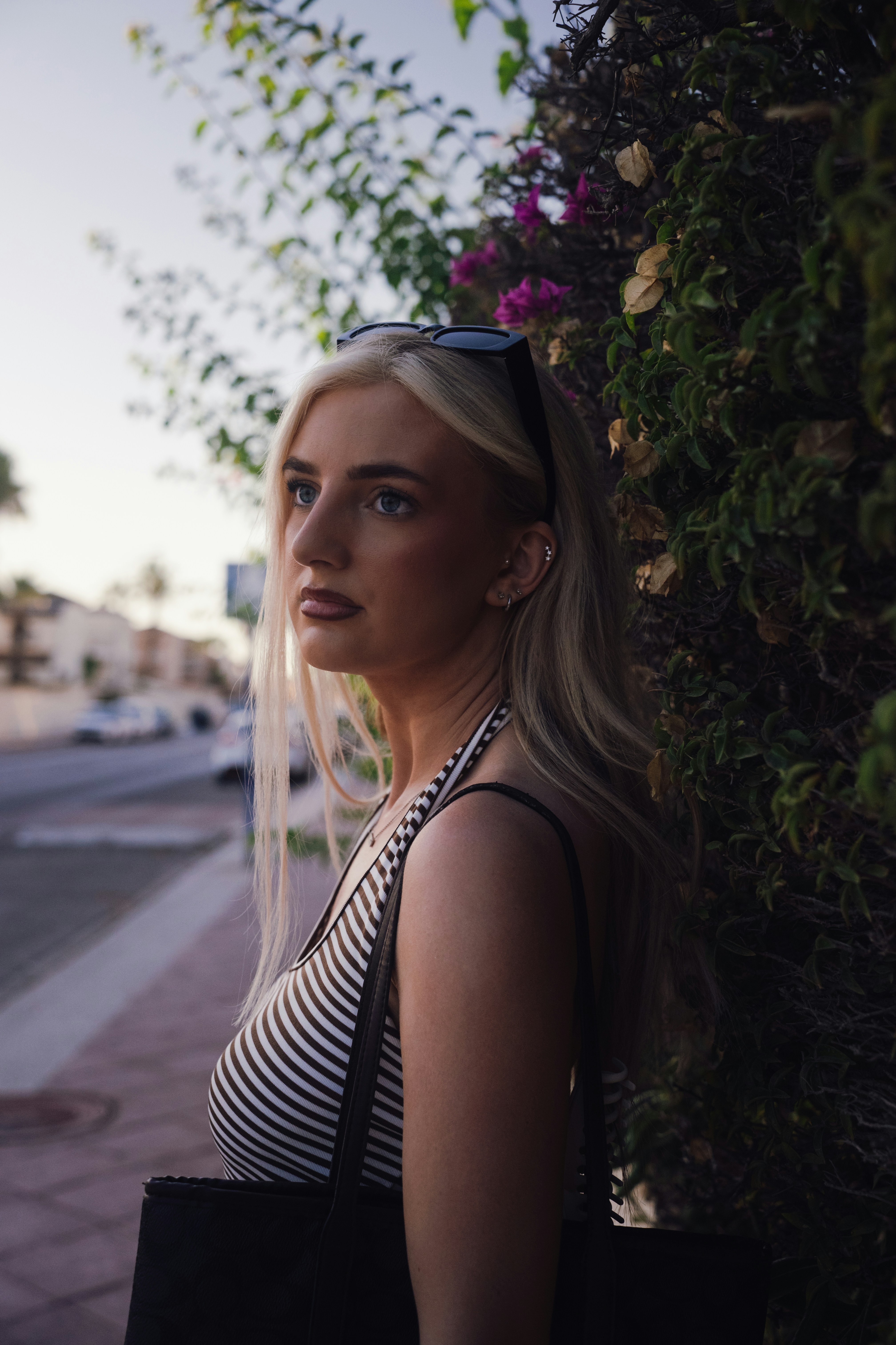 A blonde woman in a striped tank top beside a flowering hedge on a sidewalk