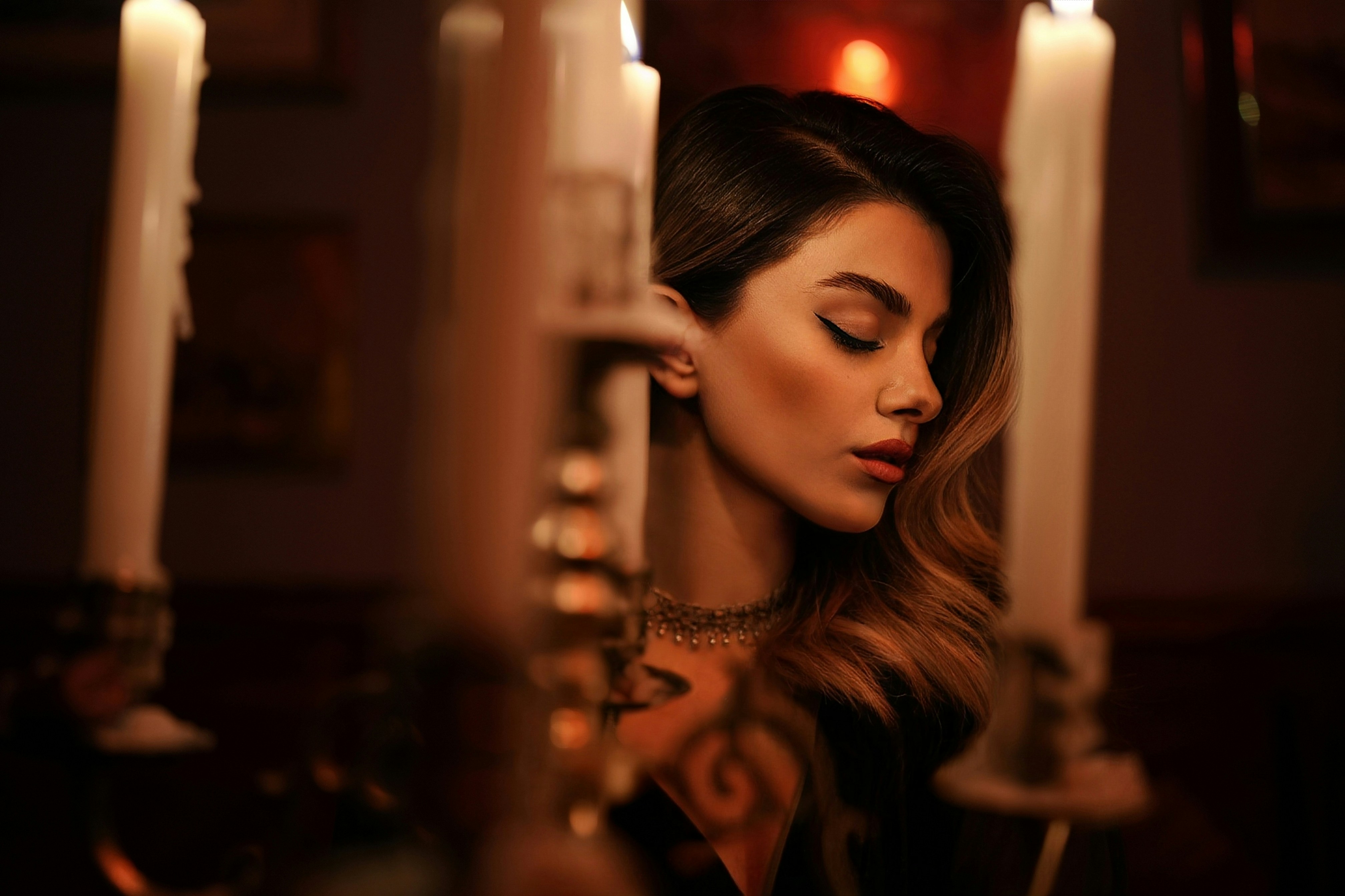 Una donna con capelli ondulati e eyeliner alato, accanto a alte candele bianche