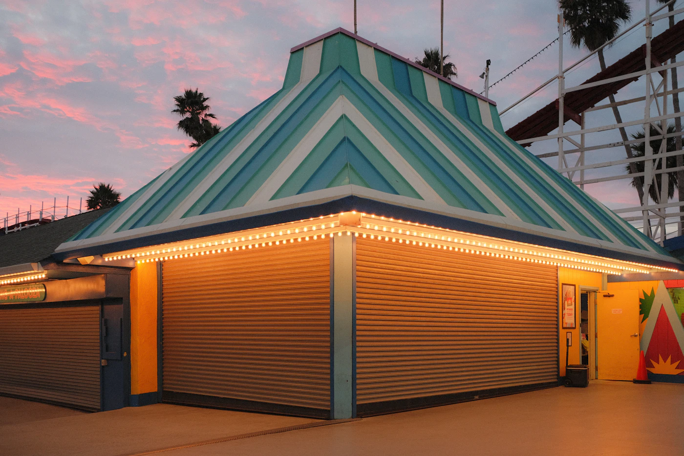 Taquilla cerrada con techo de rayas y luces encendidas en un parque de atracciones, con palmeras al atardecer