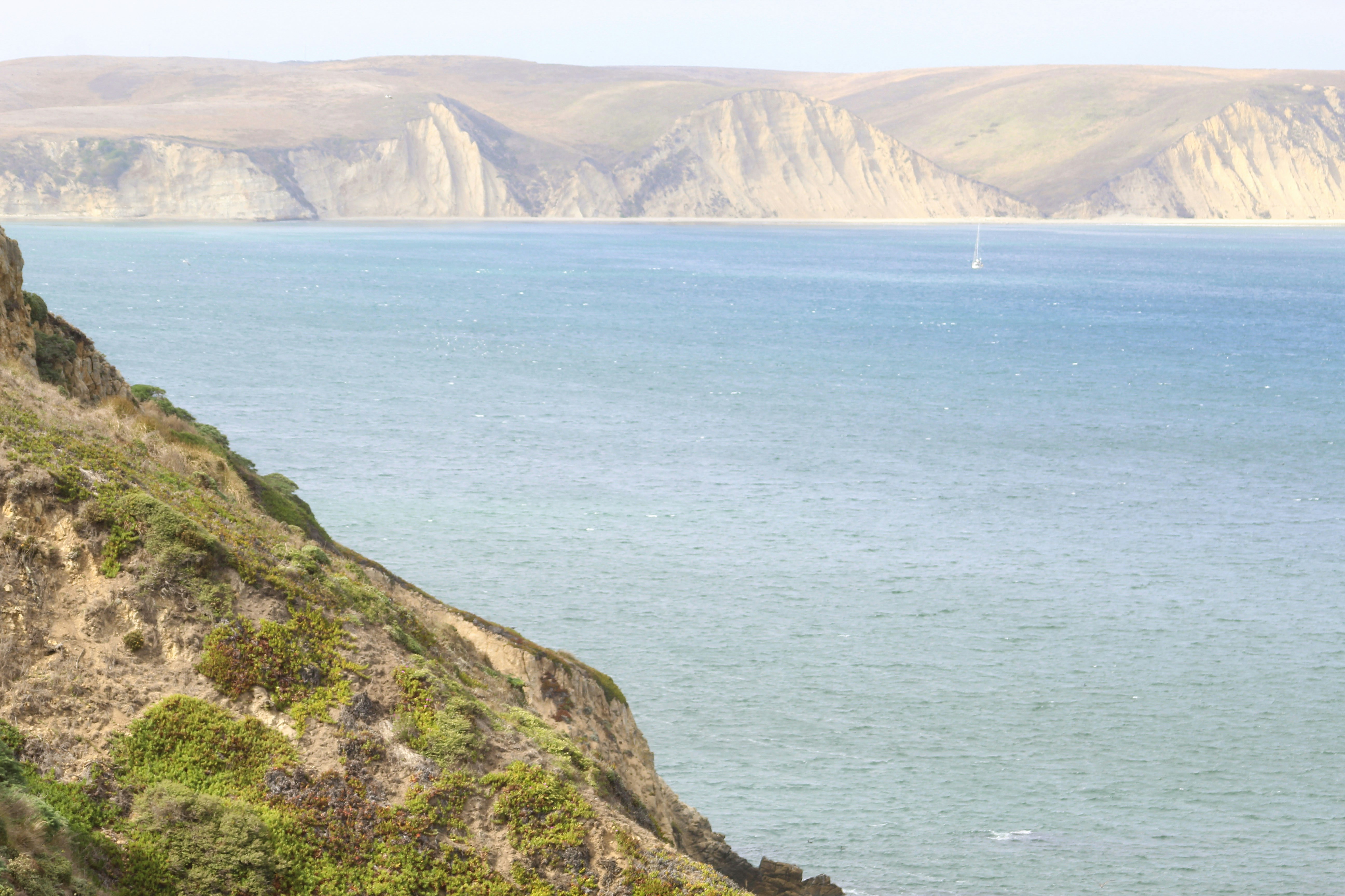 A sailboat on blue water near steep coastal cliffs and sandy hills