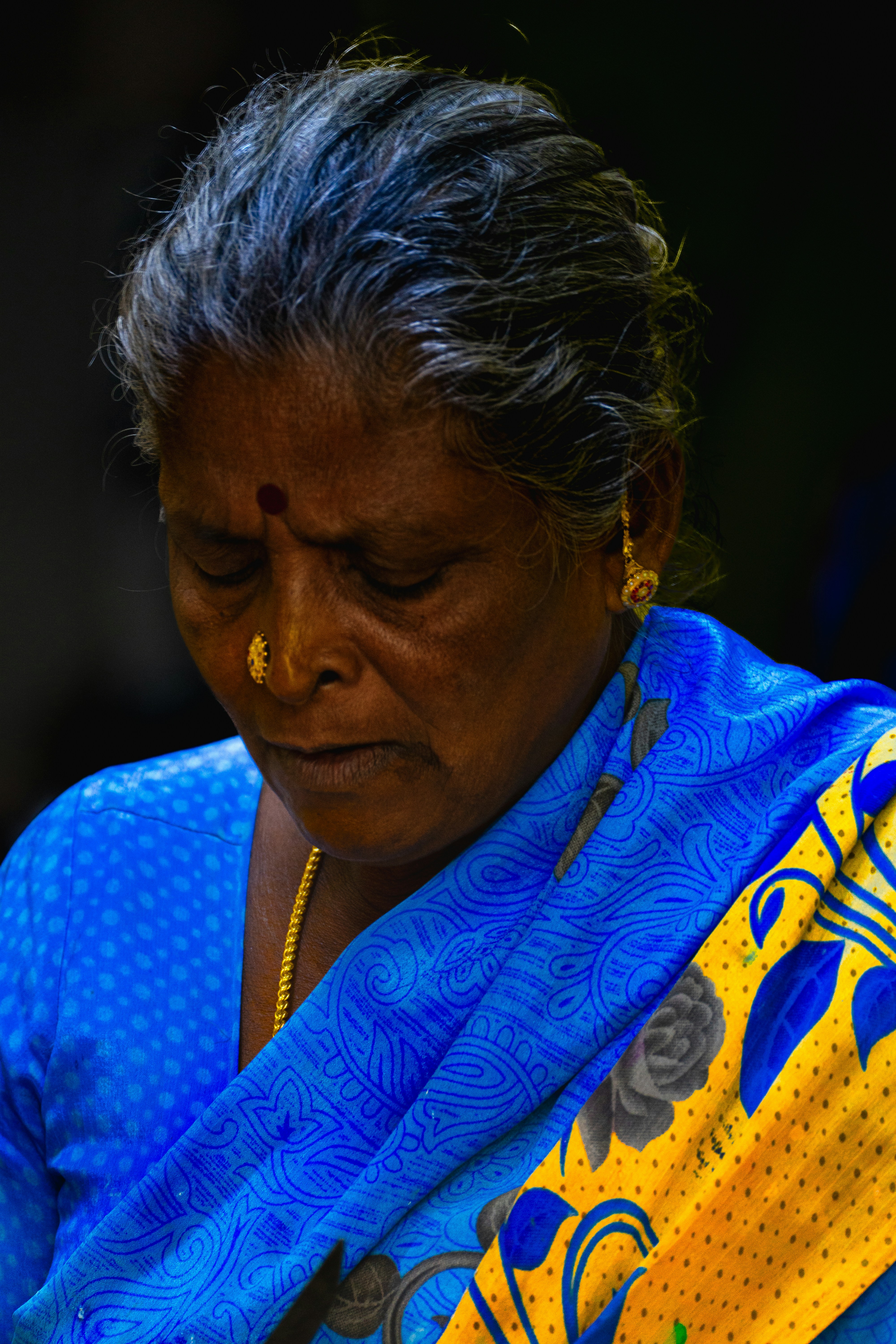 An older woman with grey hair wearing a blue and yellow patterned sari