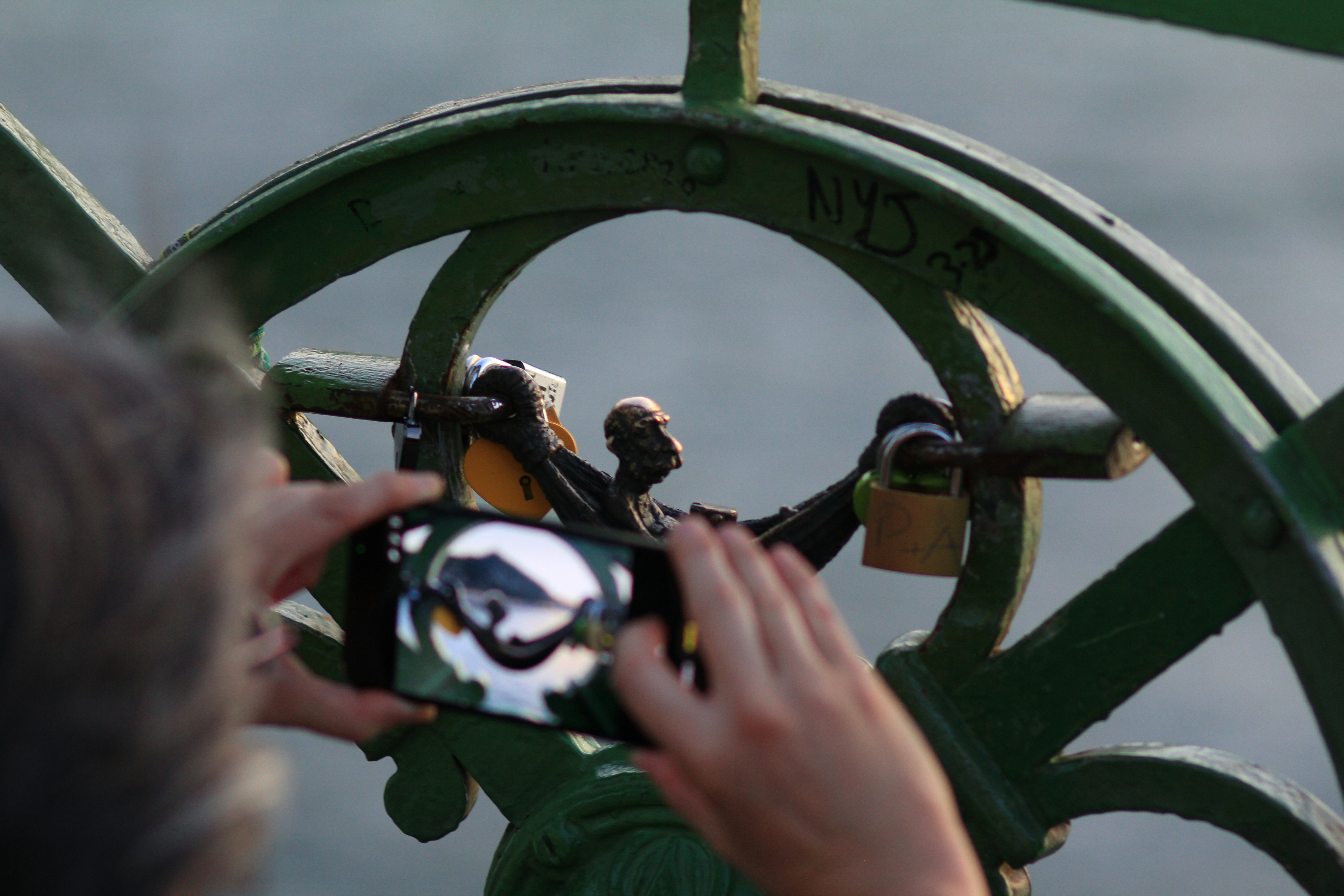 A person photographing a small bronze statue and padlocks on a green metal railing