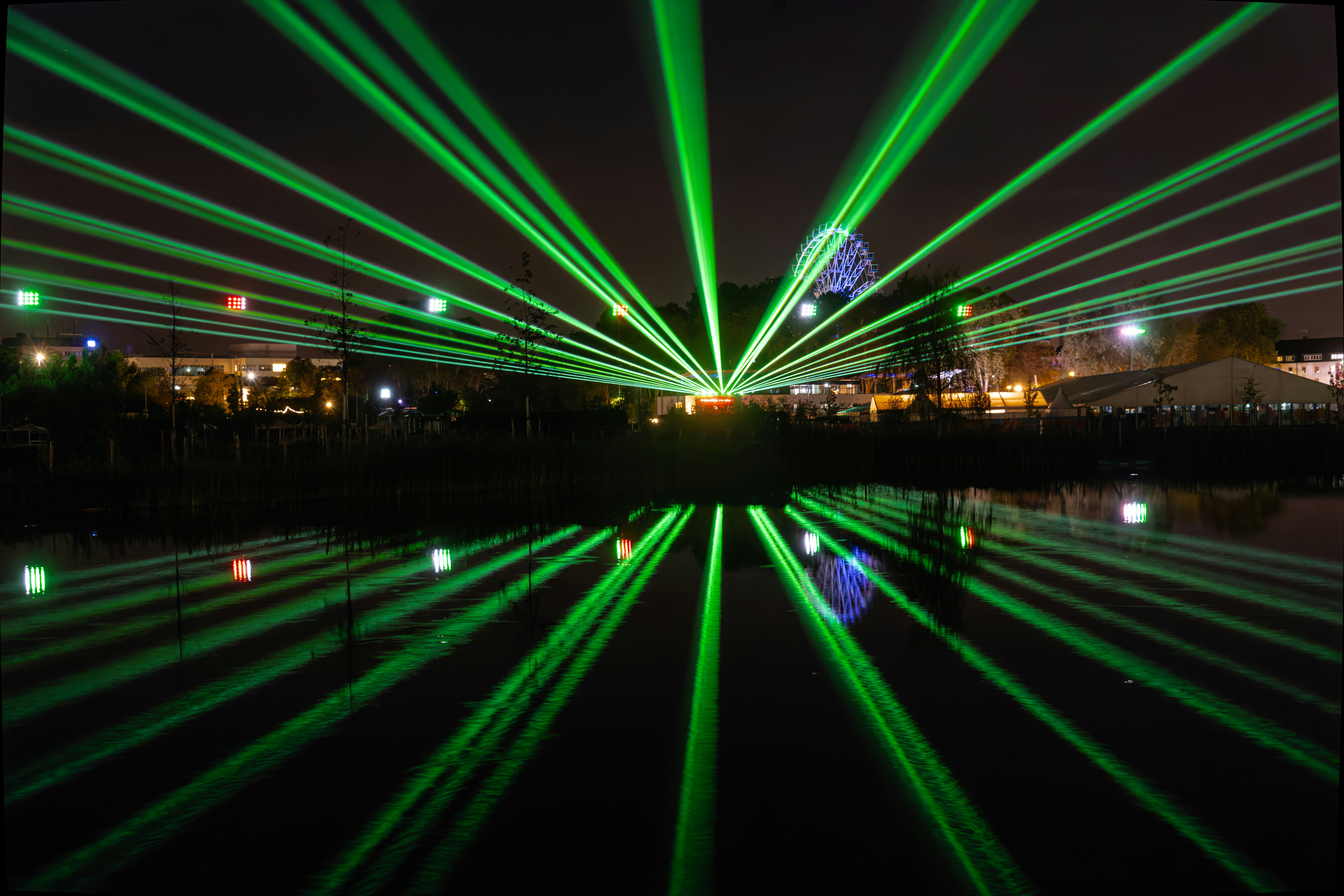 Bright green laser beams radiating over a dark lake with a ferris wheel