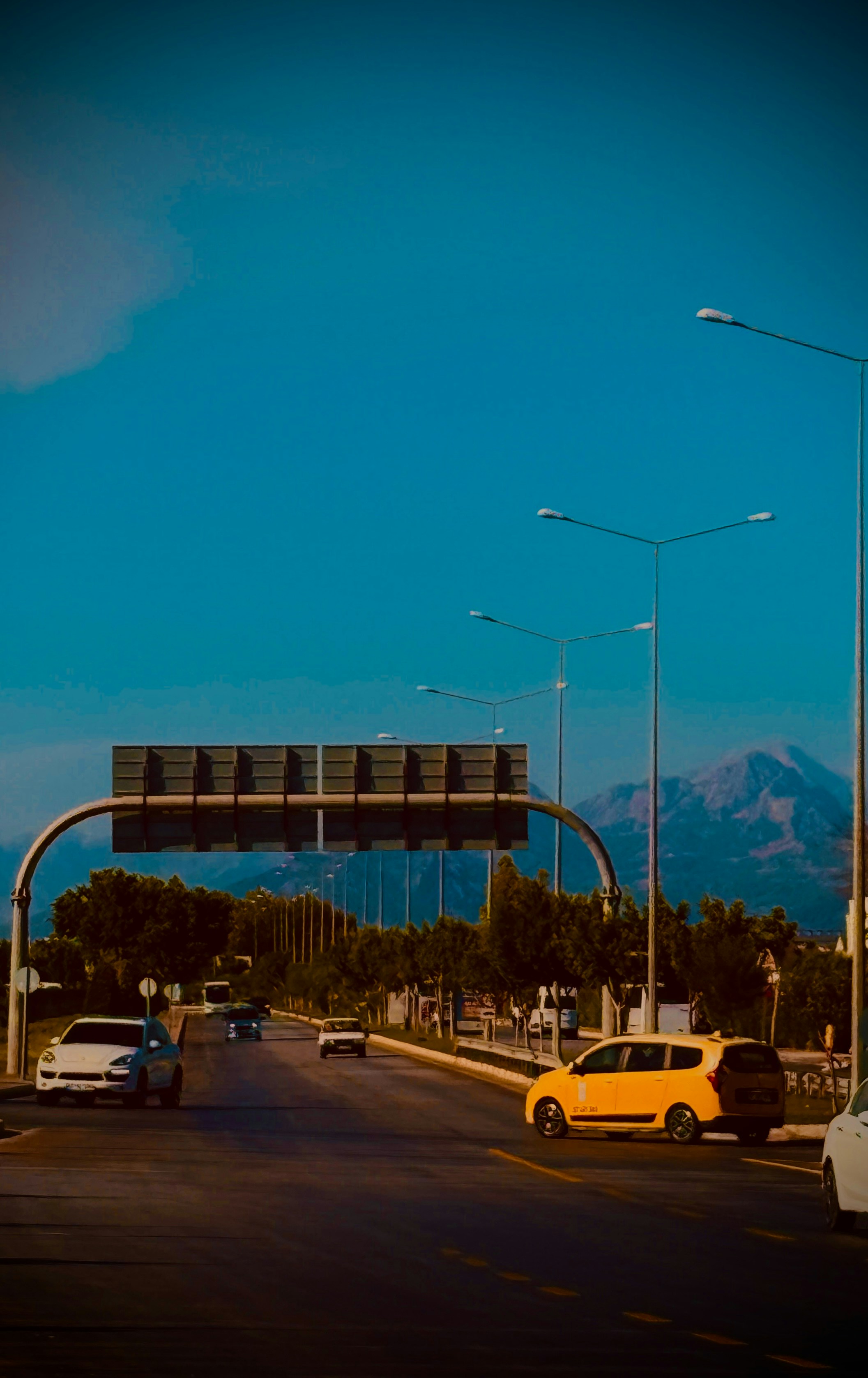 Cars driving on a highway beneath a large overhead sign with mountains in distance