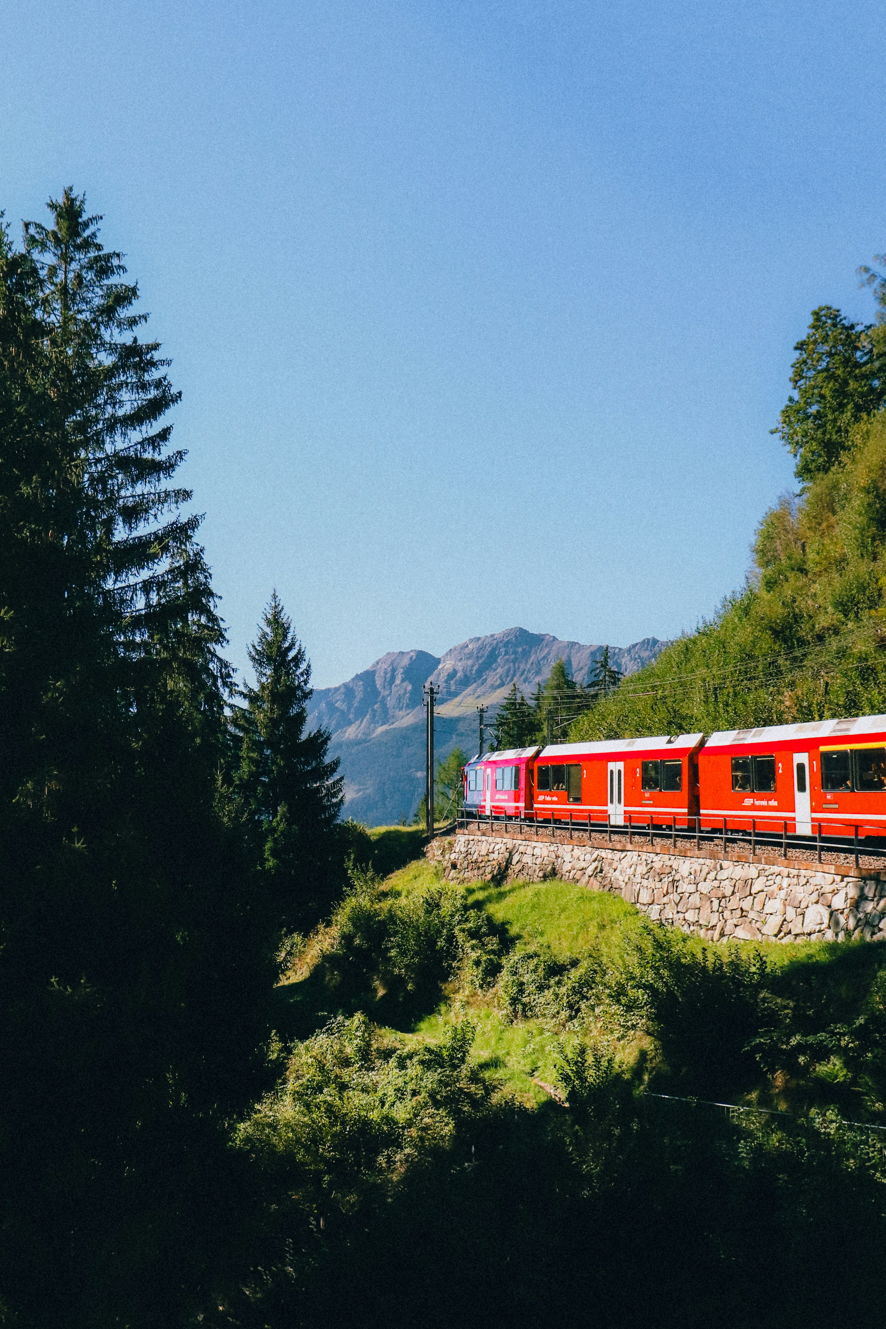A red train traveling on tracks along a stone wall in the mountains