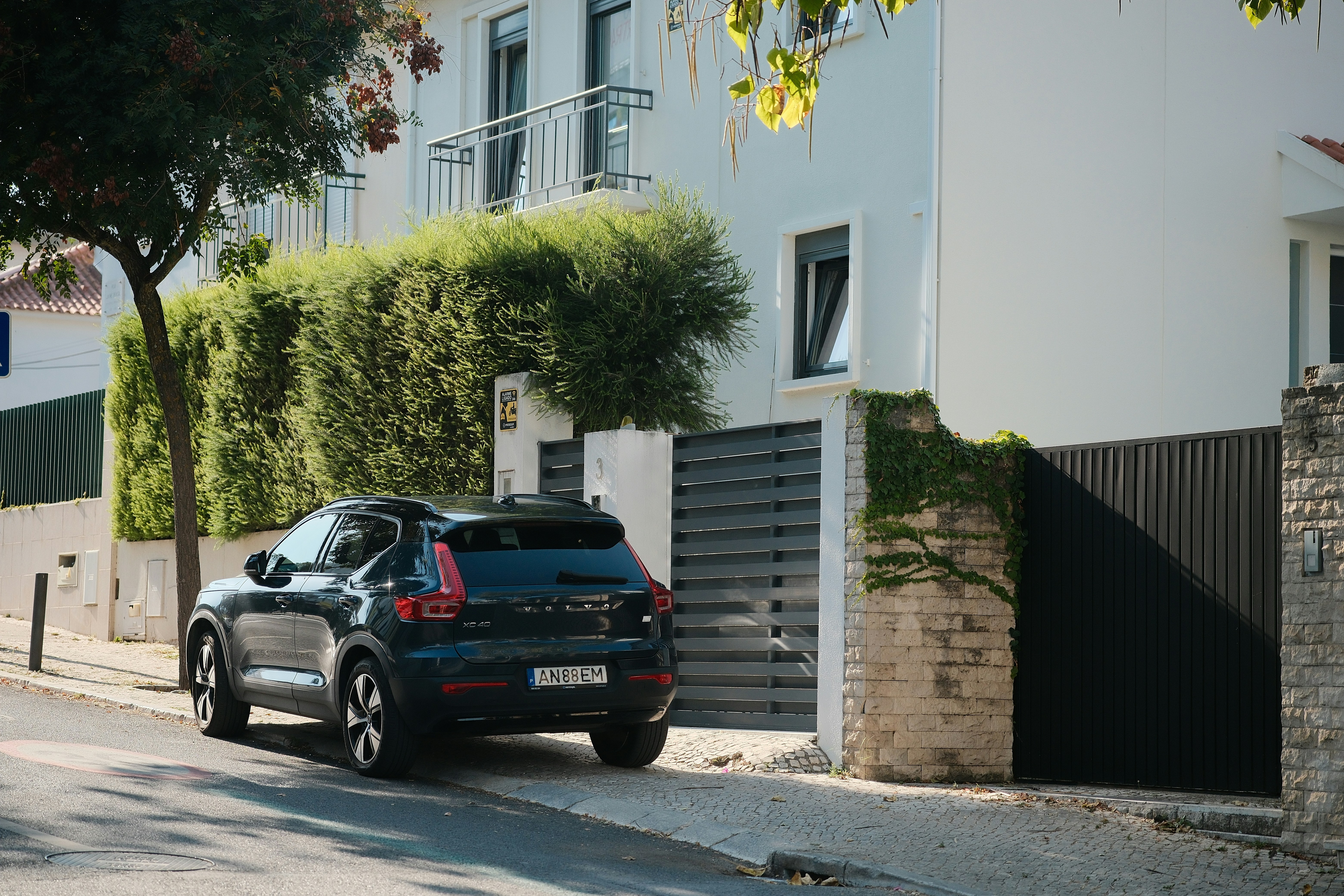 A dark Volvo XC40 parked on a street beside a white house with hedges