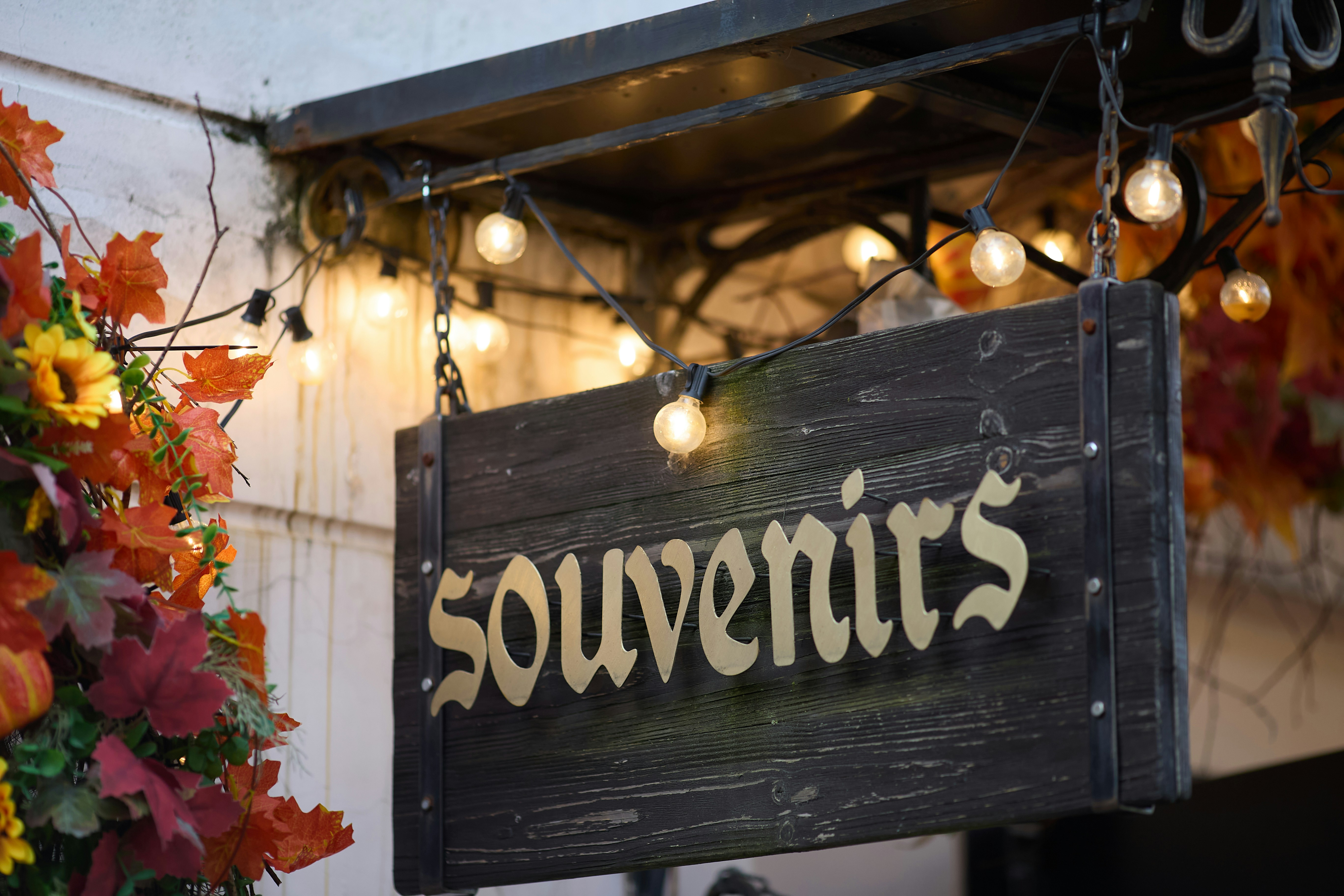 A wooden sign reading SOUVENIRS hanging under string lights with autumn leaves