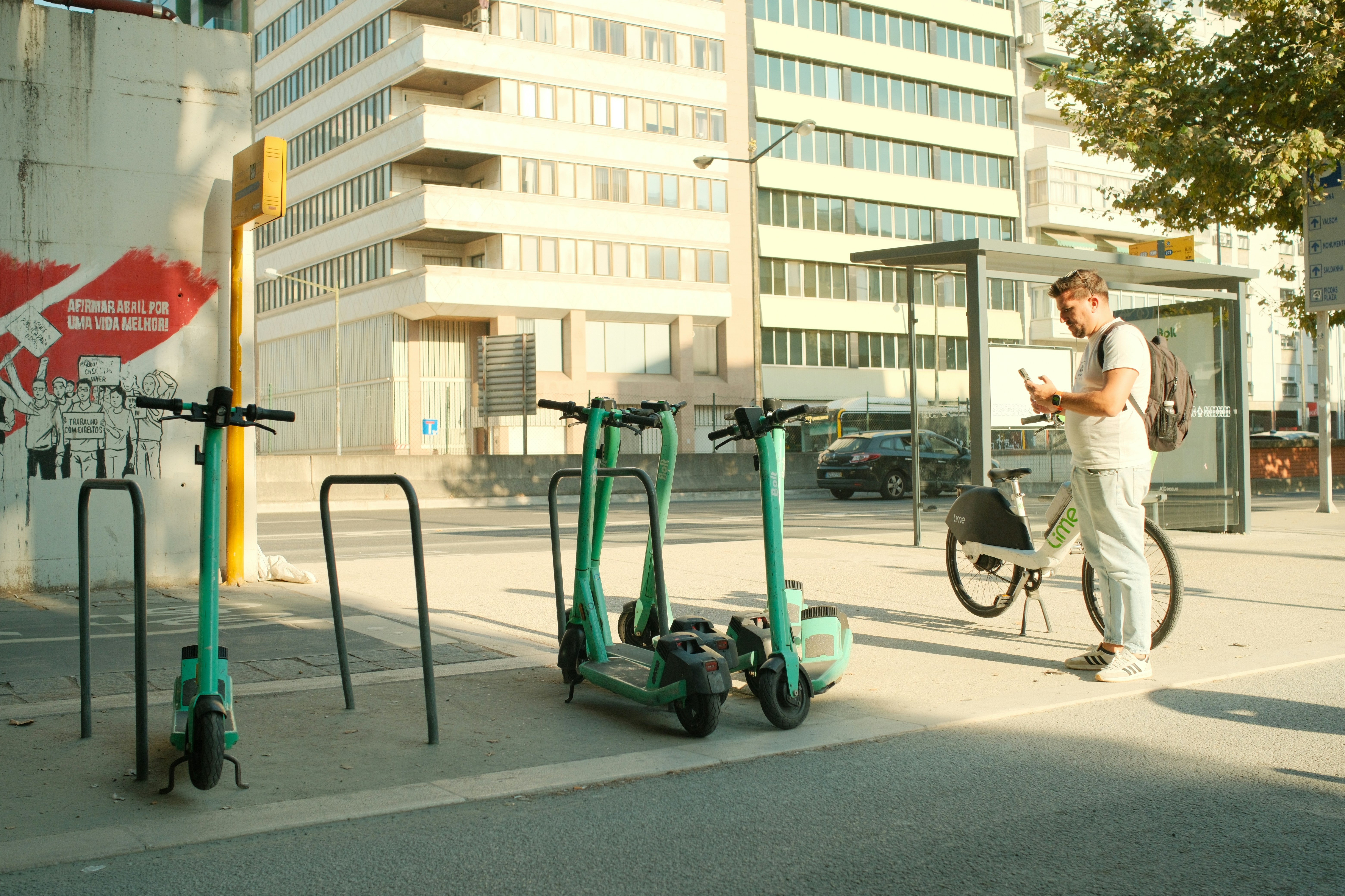 A man with a backpack using his phone beside electric scooters and a bicycle
