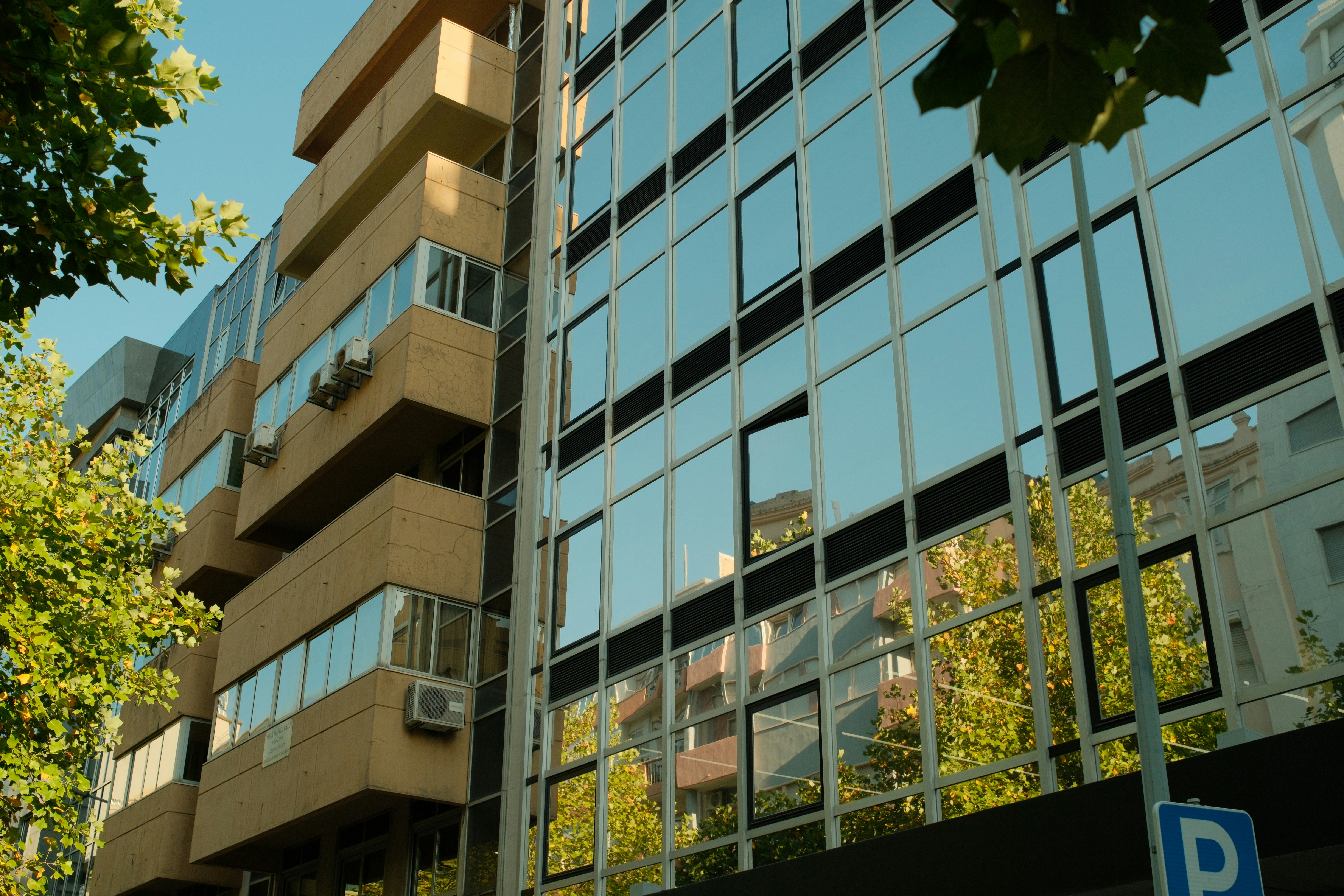 A modern glass office building reflecting green trees and nearby architecture